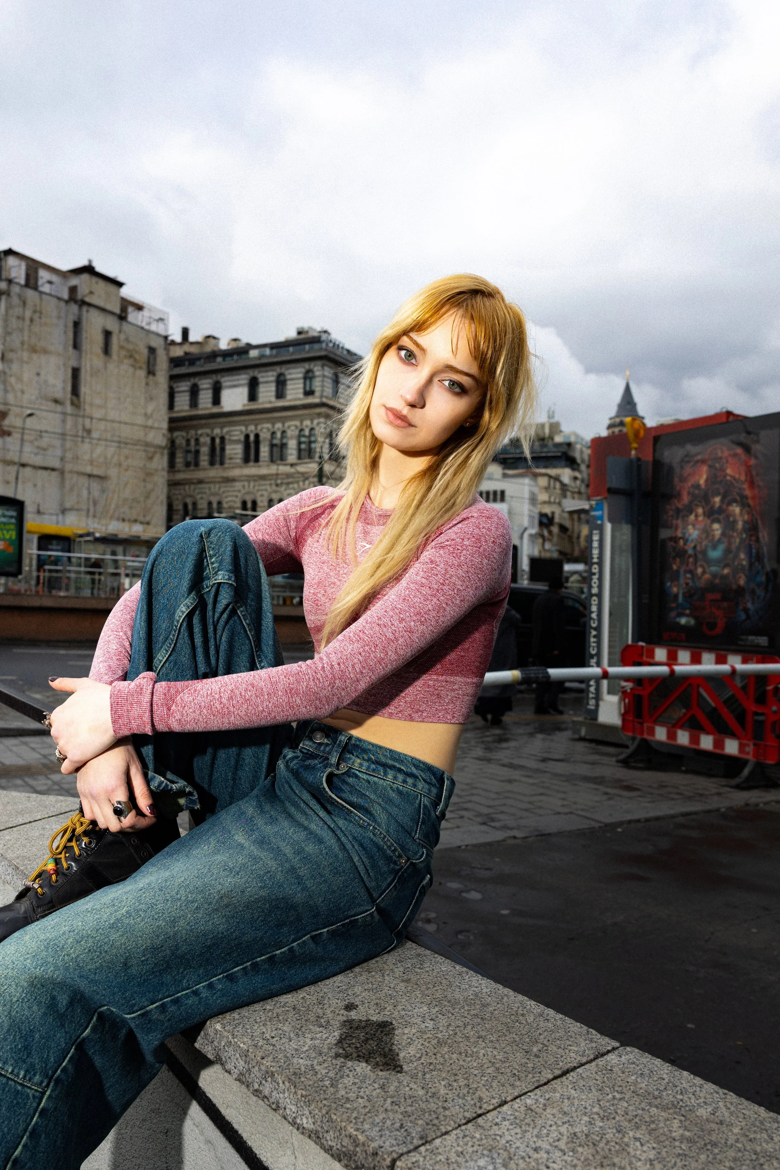 A young woman with blonde hair, wearing a pink long-sleeve top and blue jeans, sitting on a city ledge with an urban background of buildings and city life.