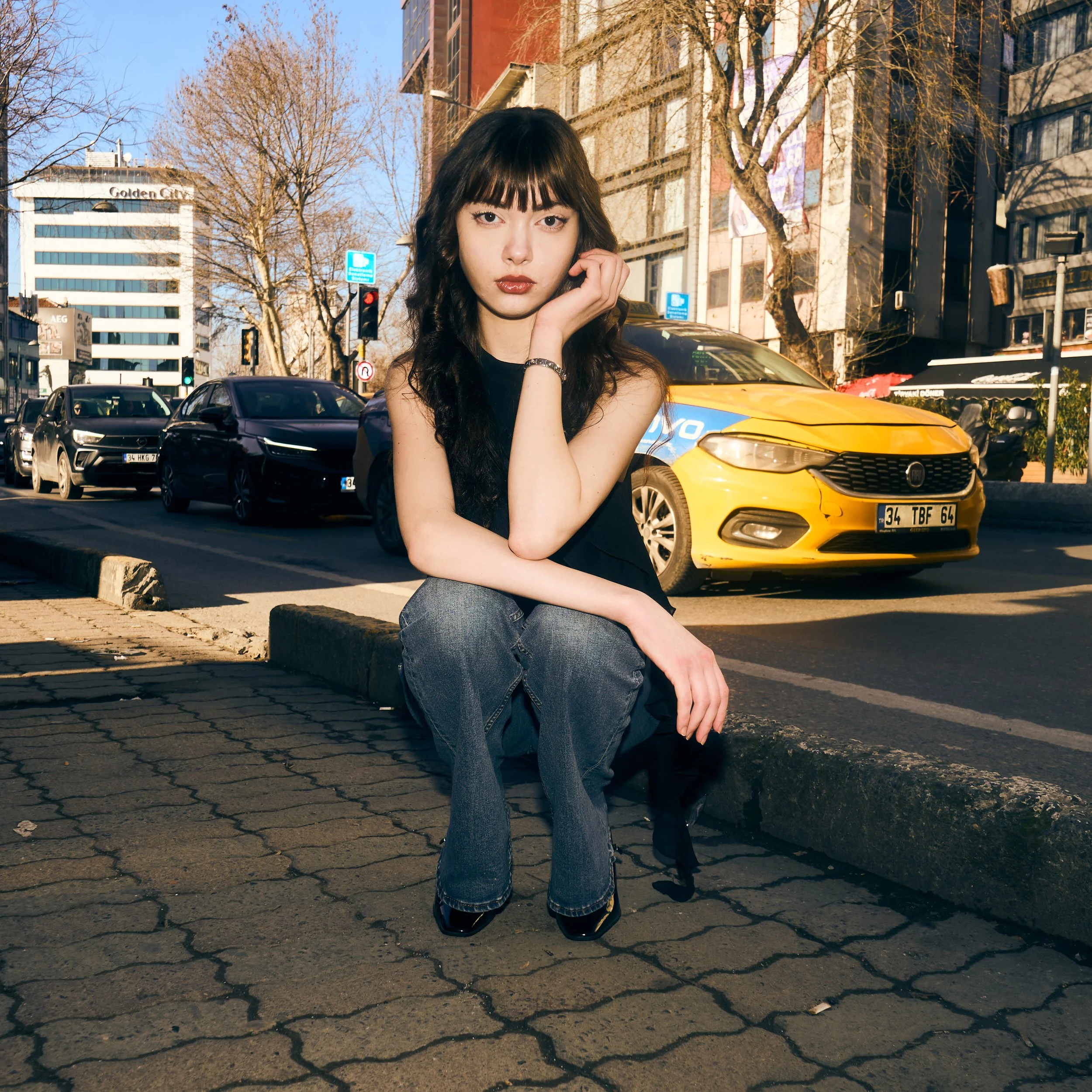 Young woman with dark hair and bangs, sitting on curb in urban street, wearing black top and jeans, with cars and tall buildings in the background.