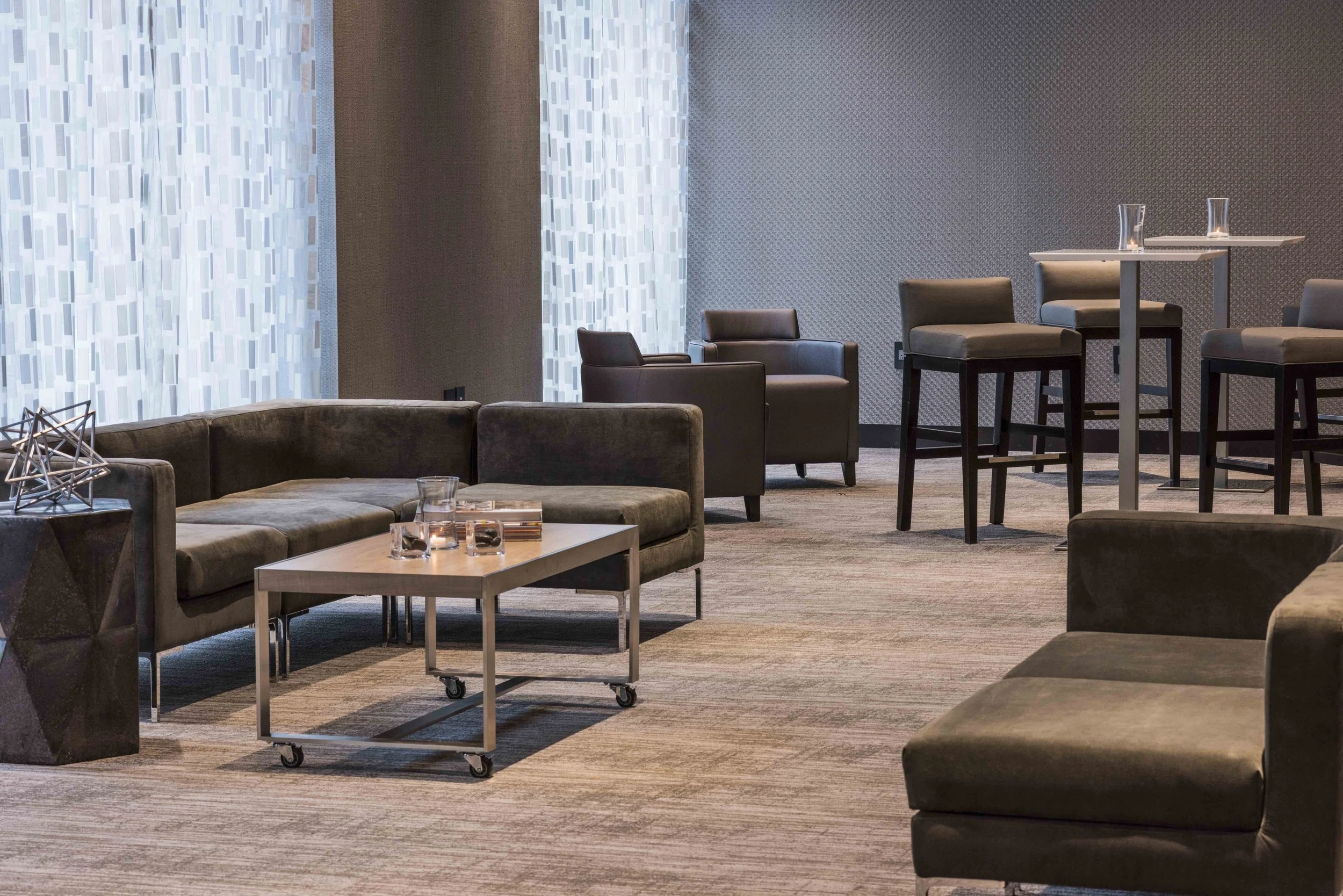 Modern lounge area with gray sofas, armchairs, and high tables with glasses, against a patterned wall and curtains.
