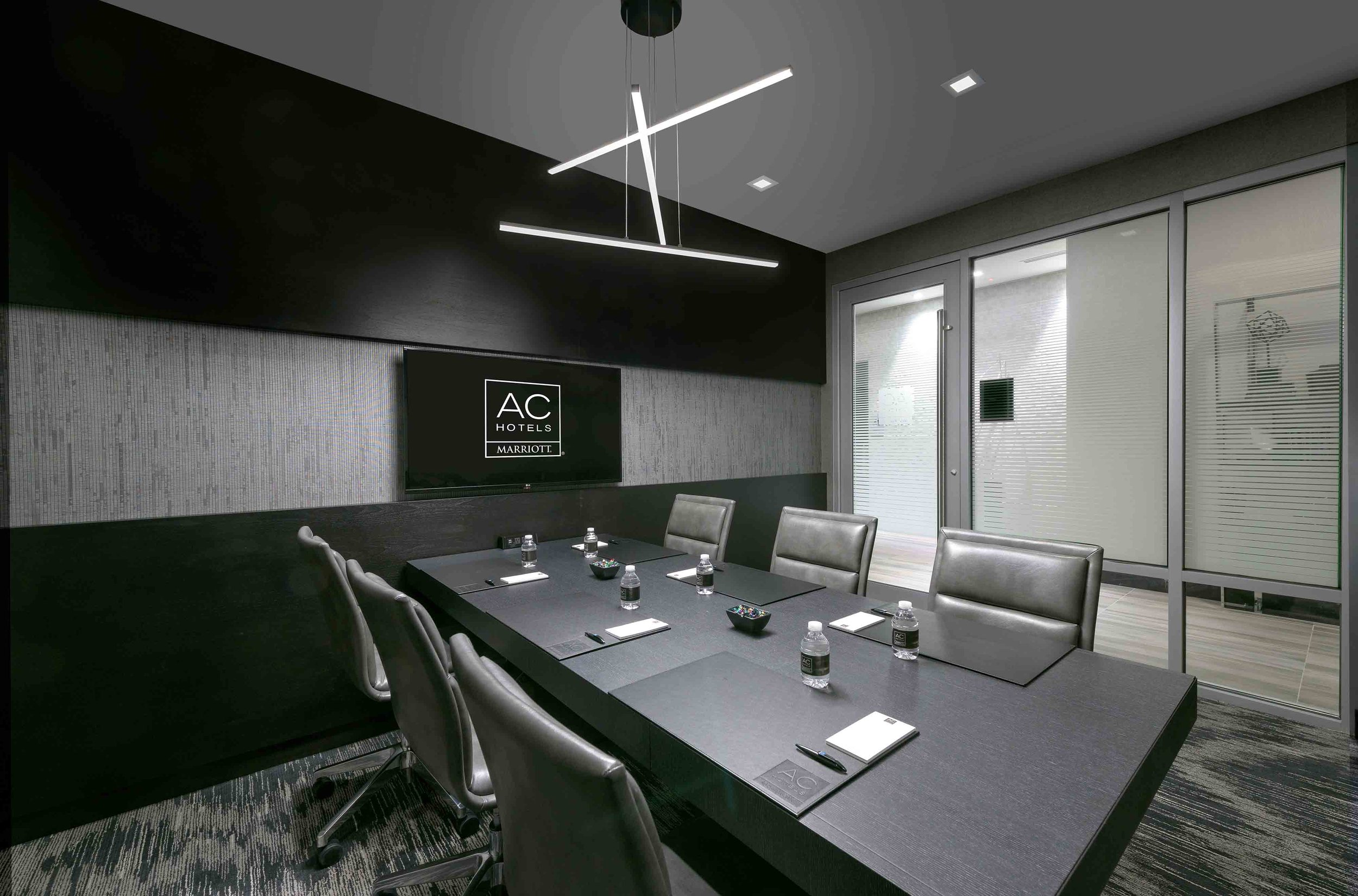 A modern conference room with a large black table, eight black leather chairs, a wall-mounted television displaying 'AC Hotels Marriott', bottled water, note pads, pens, and small bowls on the table. The room has dark gray walls, a textured gray wall panel, and large glass doors with blinds, with a contemporary pendant light fixture hanging from the ceiling.