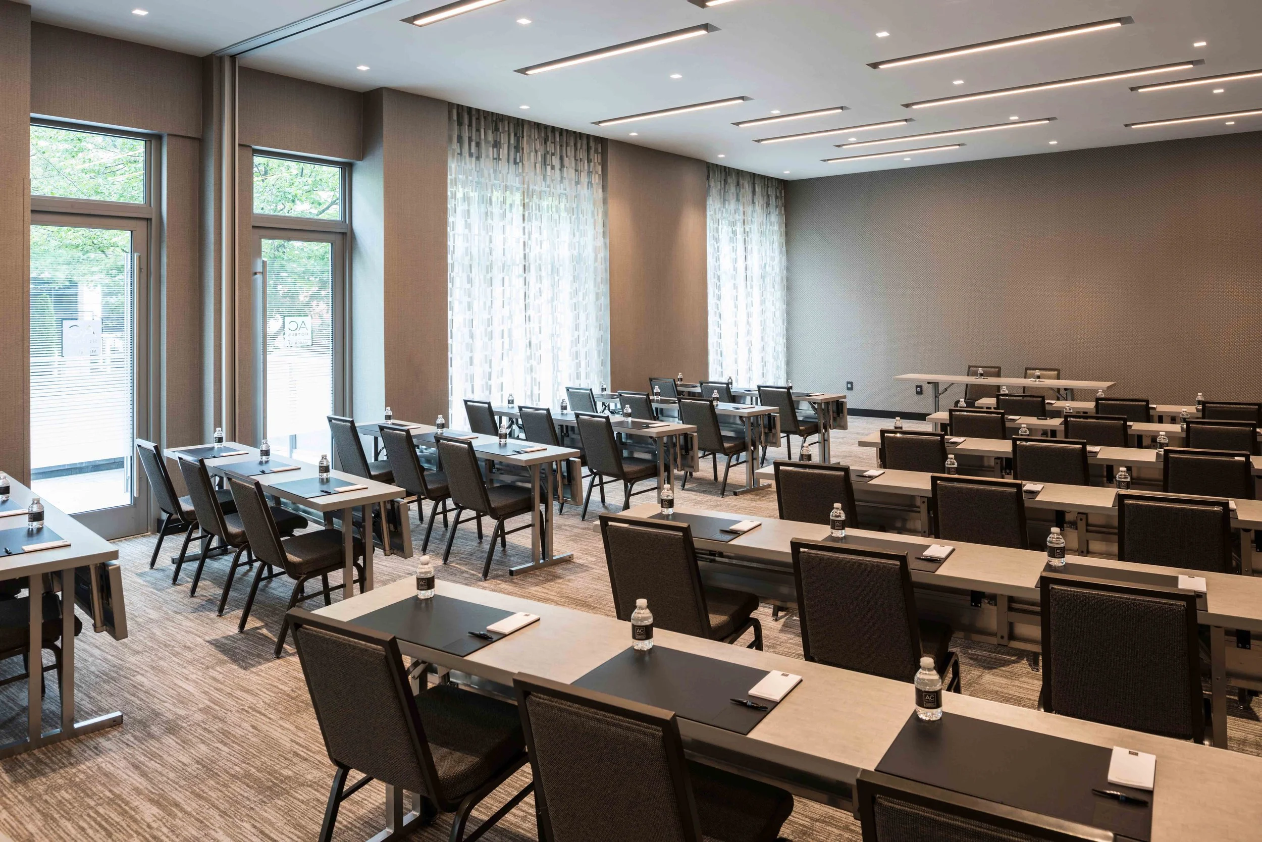Empty conference room with rows of desks, black chairs, water bottles, notepads, and pens, near large windows with curtains.