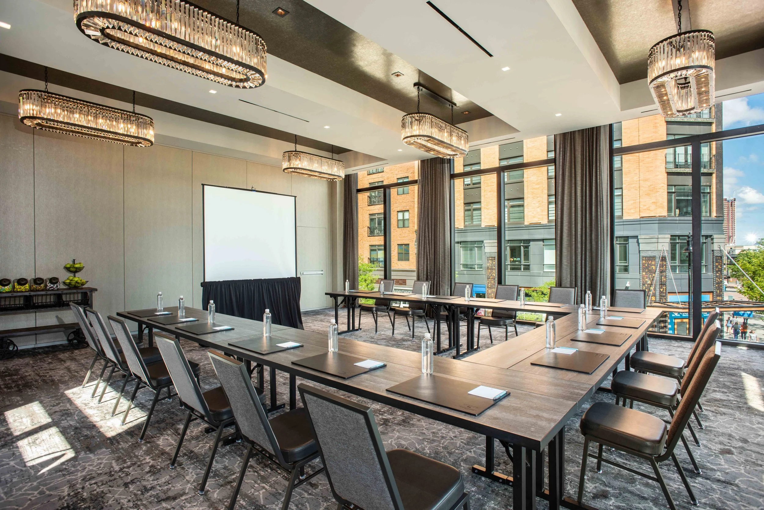 A modern conference room with U-shaped table setup, each seat has a notepad, pen, and bottle of water. There is a projector screen at the front and large windows with city views. Elegant chandeliers hang from the ceiling.