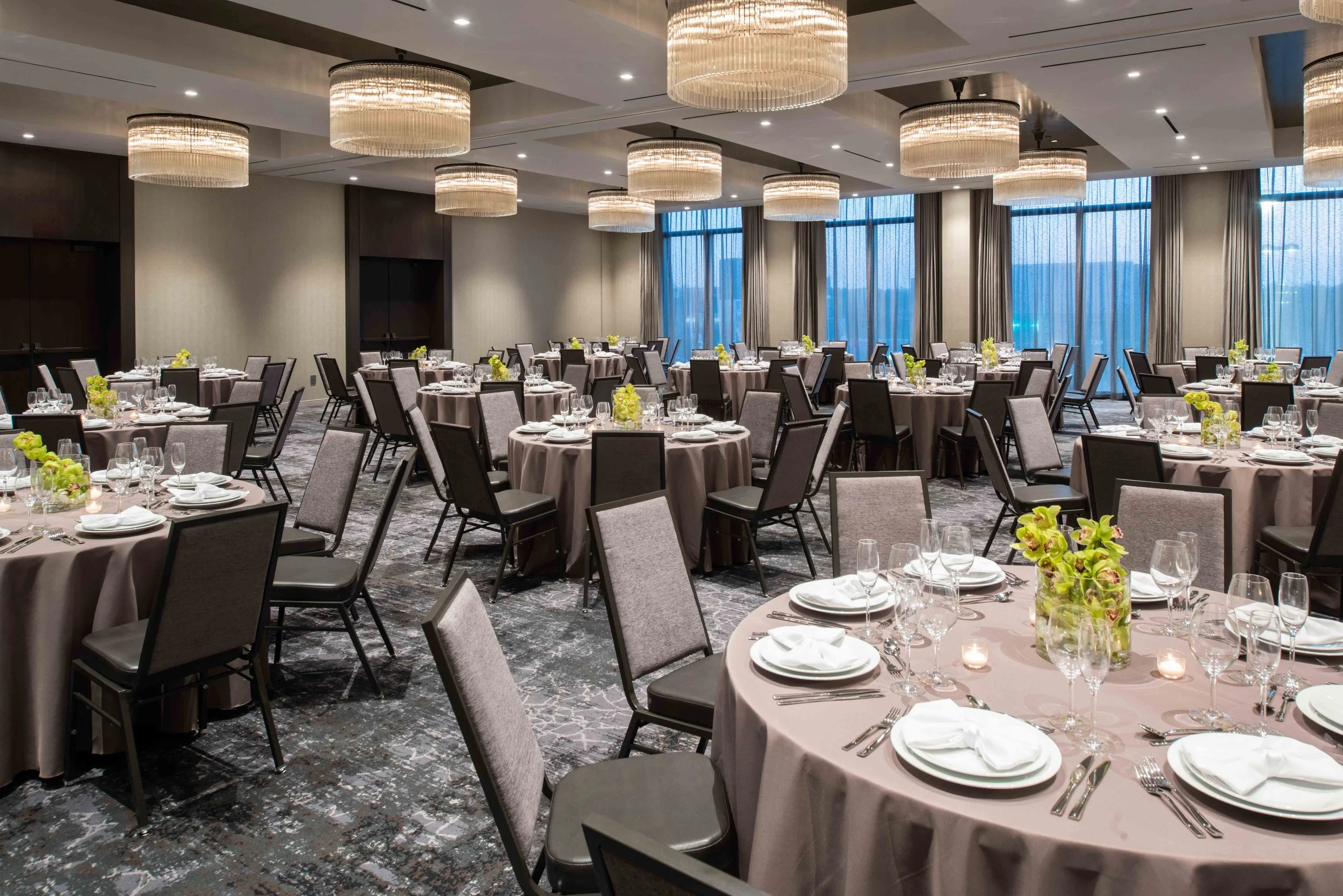 Elegant banquet hall set up for an event with round tables covered in light pink tablecloths, decorated with floral centerpieces, wine glasses, and white dinnerware. Modern chandelier lighting fixtures hang from the ceiling. Large windows with sheer curtains allow natural light to fill the room.