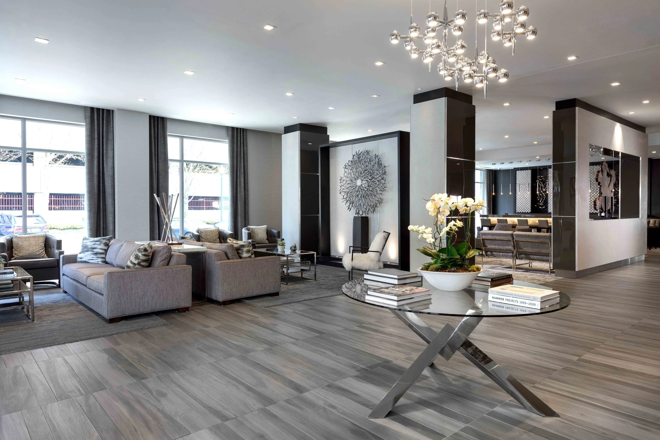 Modern spacious lobby with light gray sofas, large windows with dark curtains, a glass coffee table, a round glass table with a white orchid plant, a stylish chandelier, and decorative wall art.