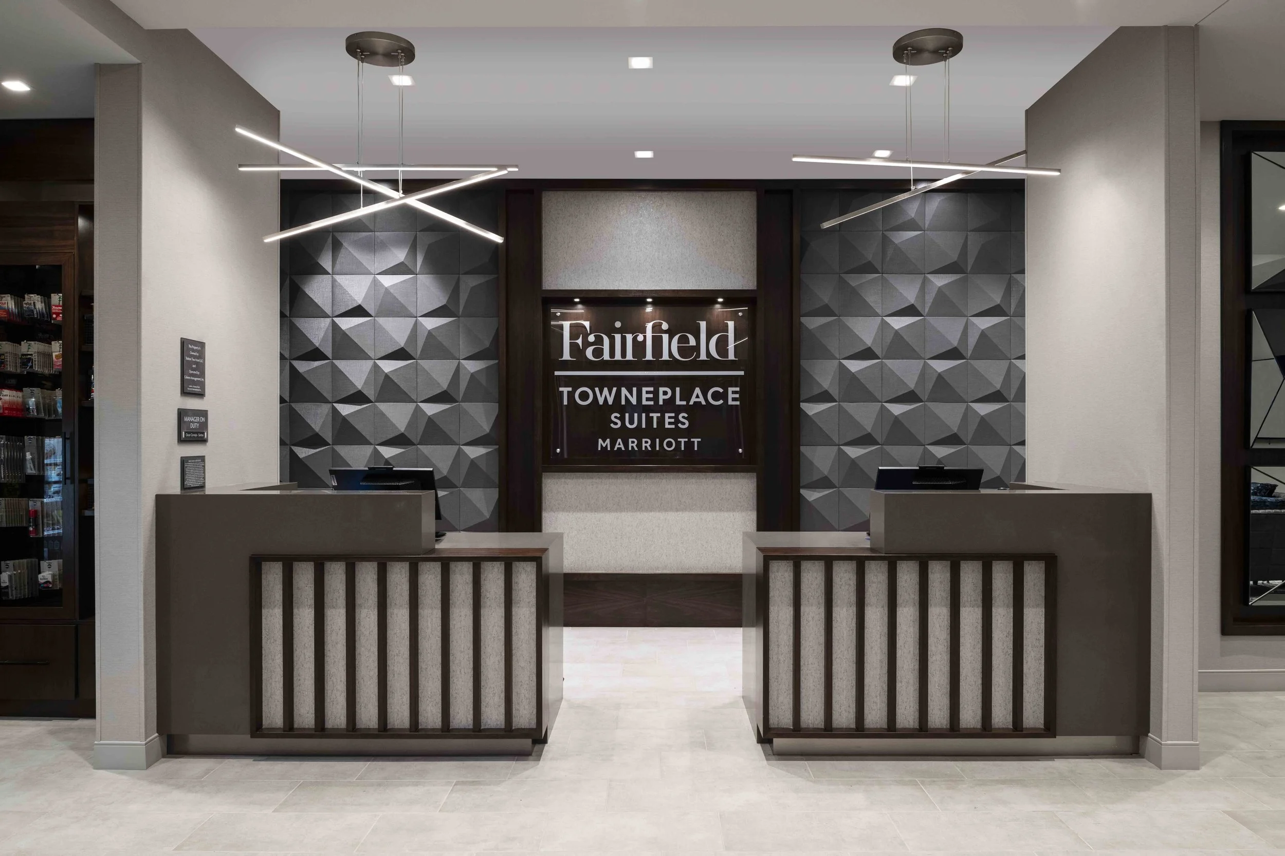 Hotel lobby reception area with two check-in counters, a decorative wall with geometric patterns, and a sign reading 'Fairfield Towneplace Suites Marriott' in the background.