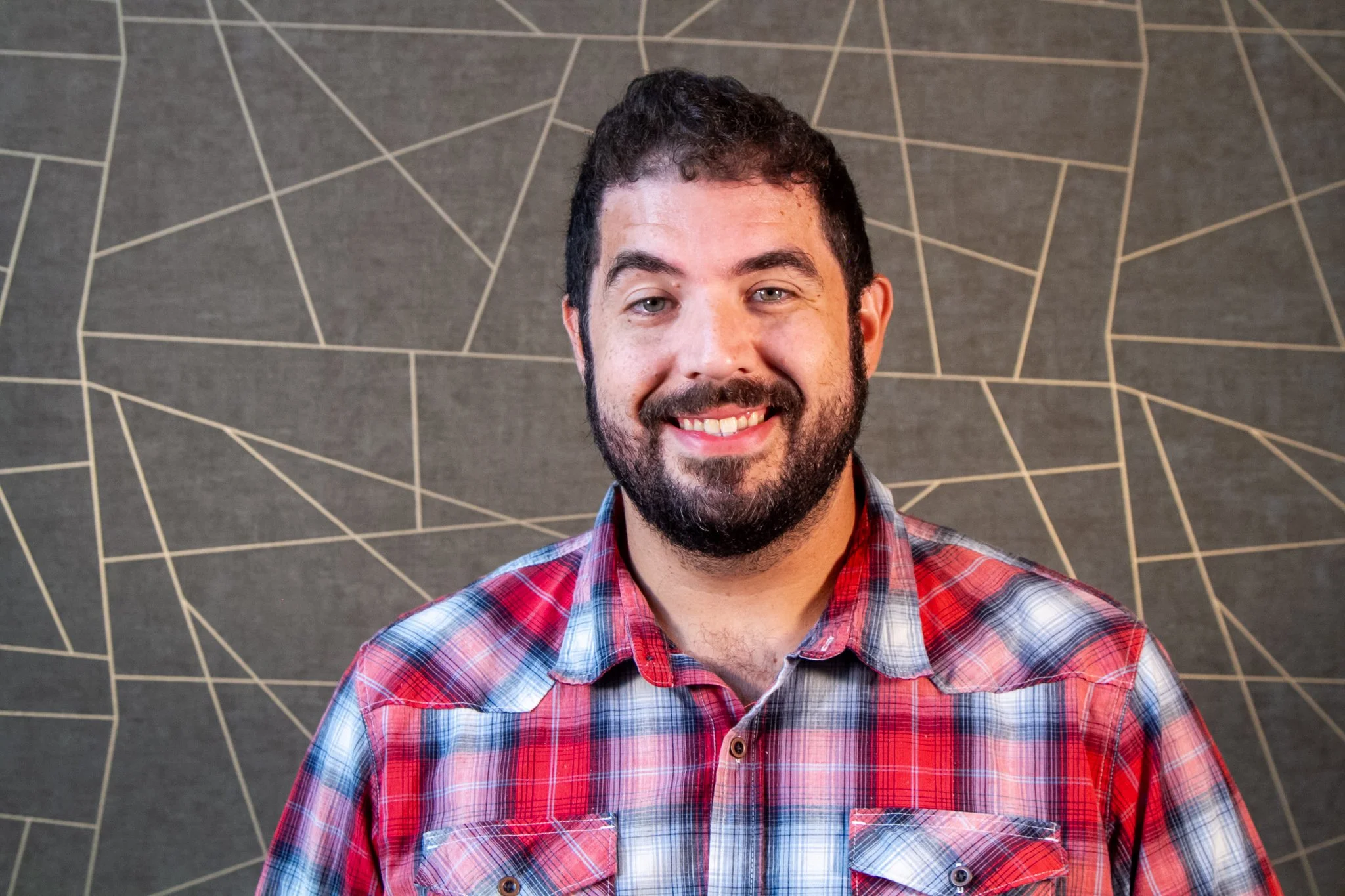 A man with a beard and dark curly hair smiling at the camera, wearing a red and gray plaid shirt, standing in front of a wall with gray geometric patterns.