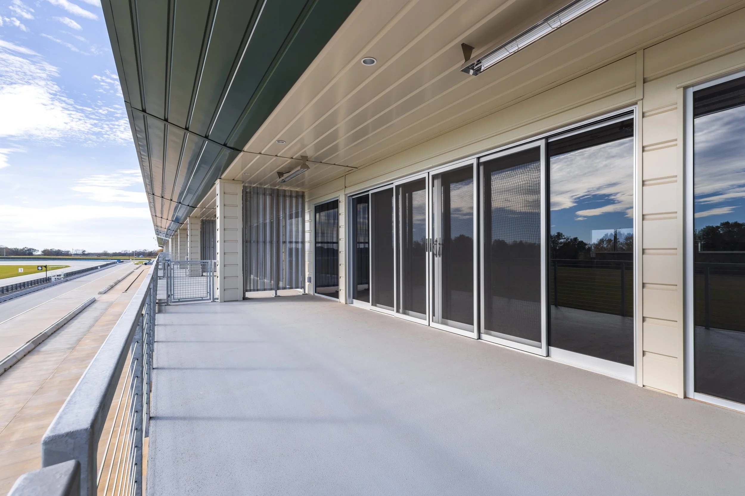 Empty balcony with sliding glass doors and black mesh screens, overlooking a racetrack and grassy fields under a partly cloudy sky.