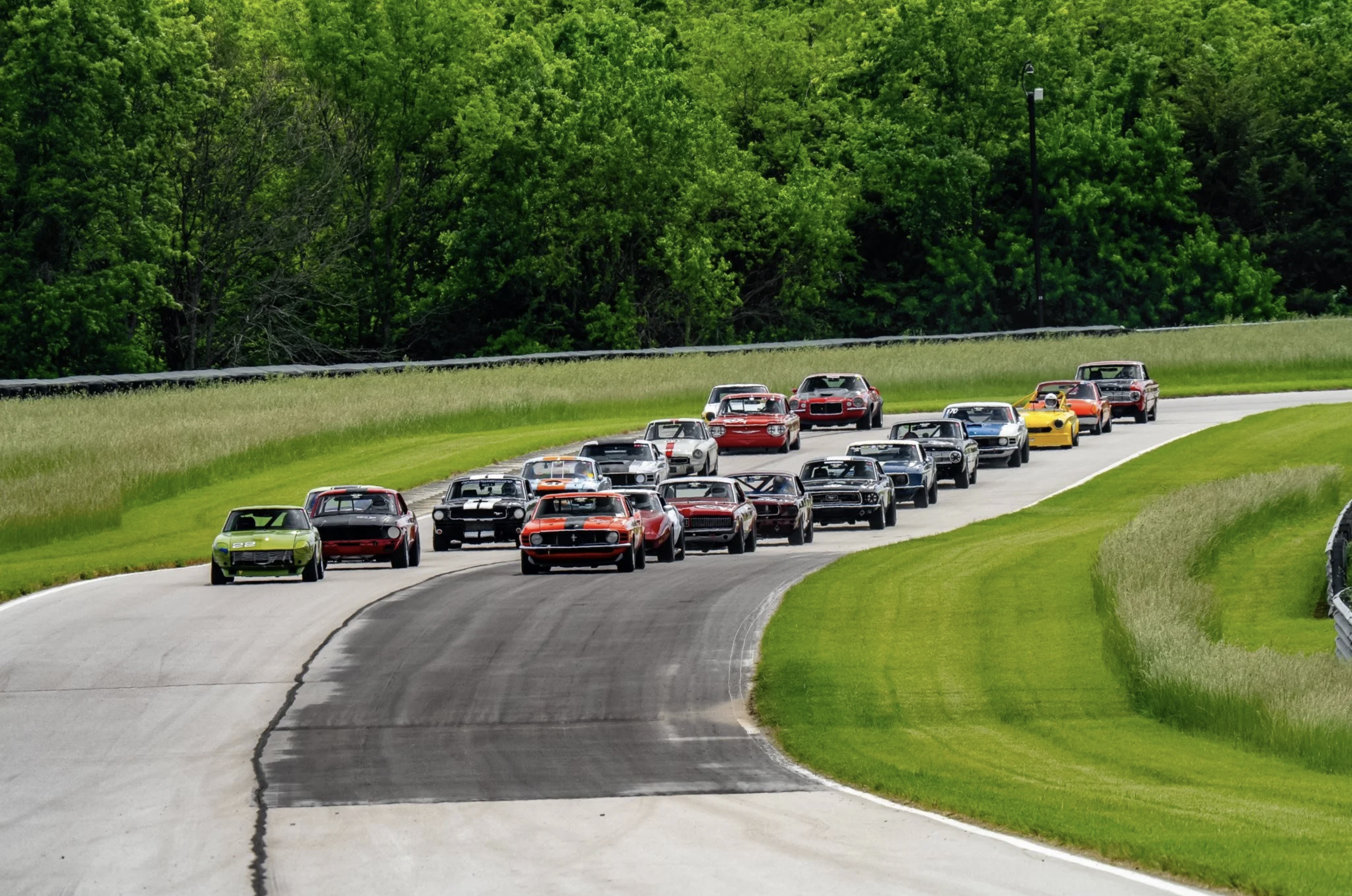 A group of race cars on a race track, surrounded by green grass and trees.