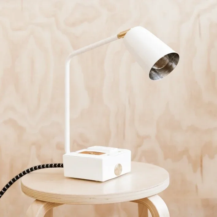 A white adjustable desk lamp with a brass accent, placed on a round wooden side table against a light-colored wall.