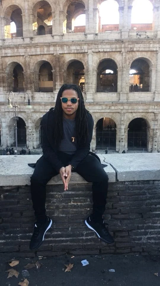 A man with long dreadlocks wearing sunglasses, a dark jacket, and black pants sitting on a stone wall in front of the Colosseum in Rome, Italy.