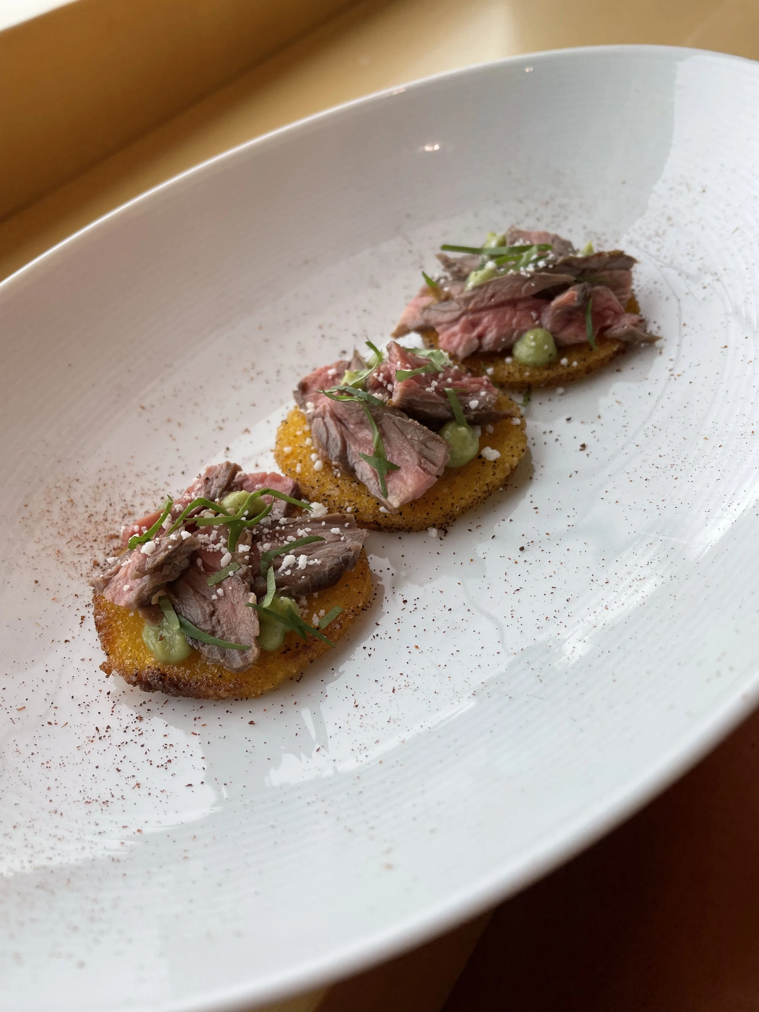 Three pieces of crostini topped with sliced beef, green purée, and garnished with herbs on a white plate.
