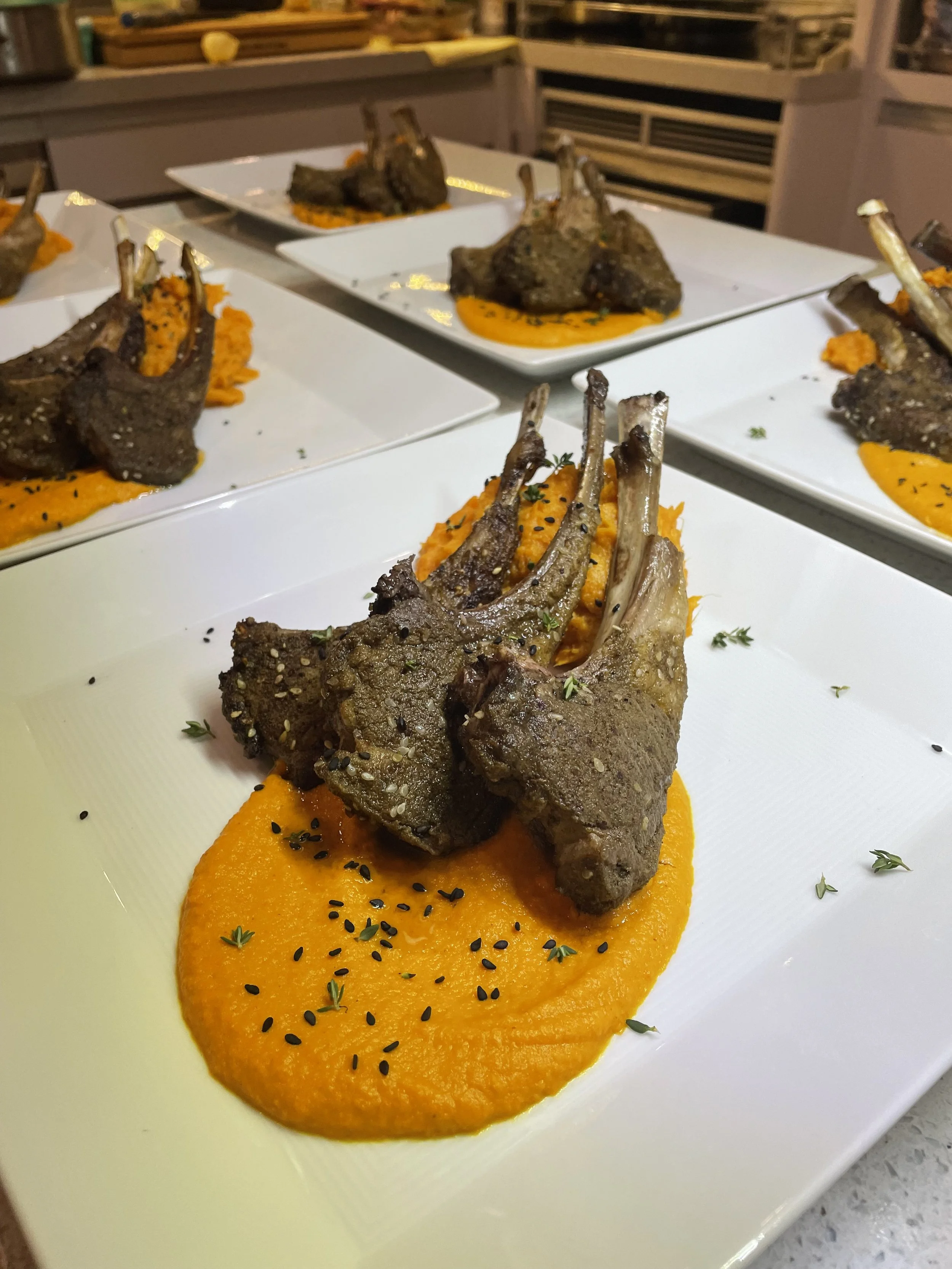 Plated lamb chops served on a bed of orange mashed sweet potatoes, garnished with black sesame seeds and herbs.
