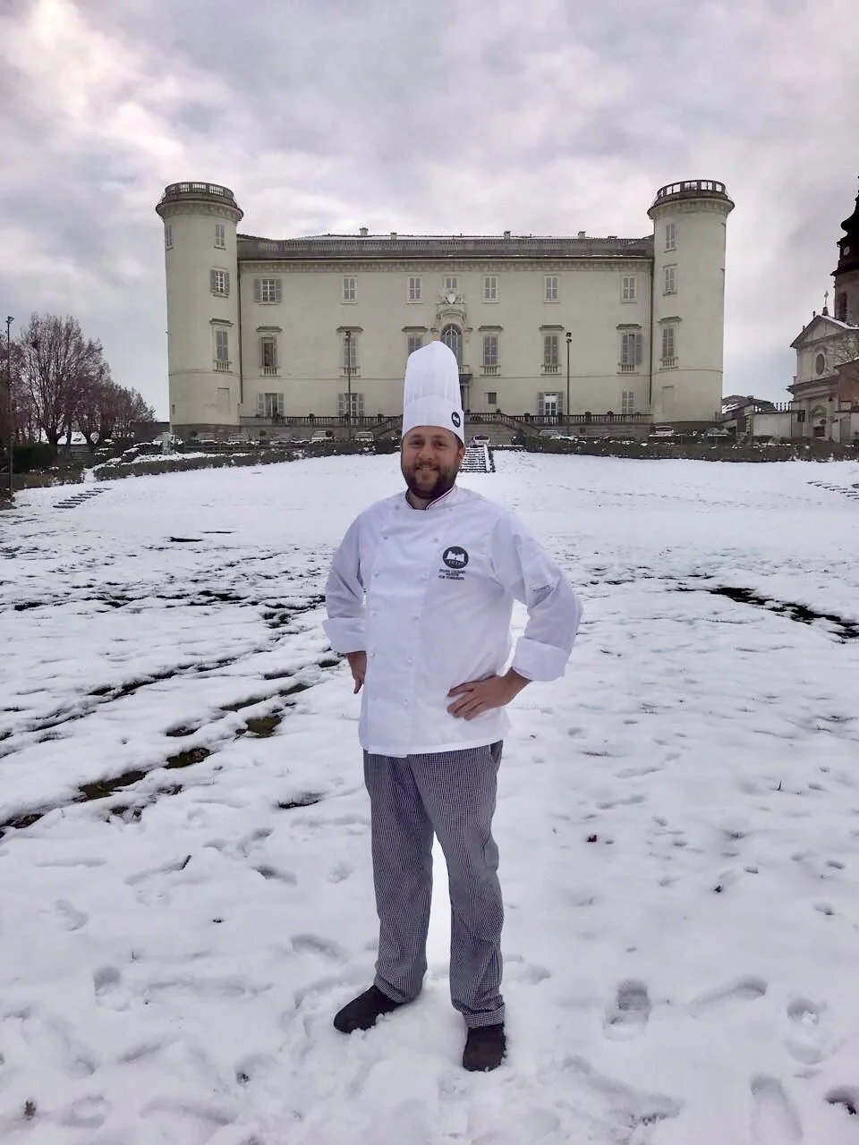 A man dressed as a chef in a white chef's coat and tall hat, standing outdoors in the snow in front of a large historic building, possibly a castle or mansion, on a cloudy day.