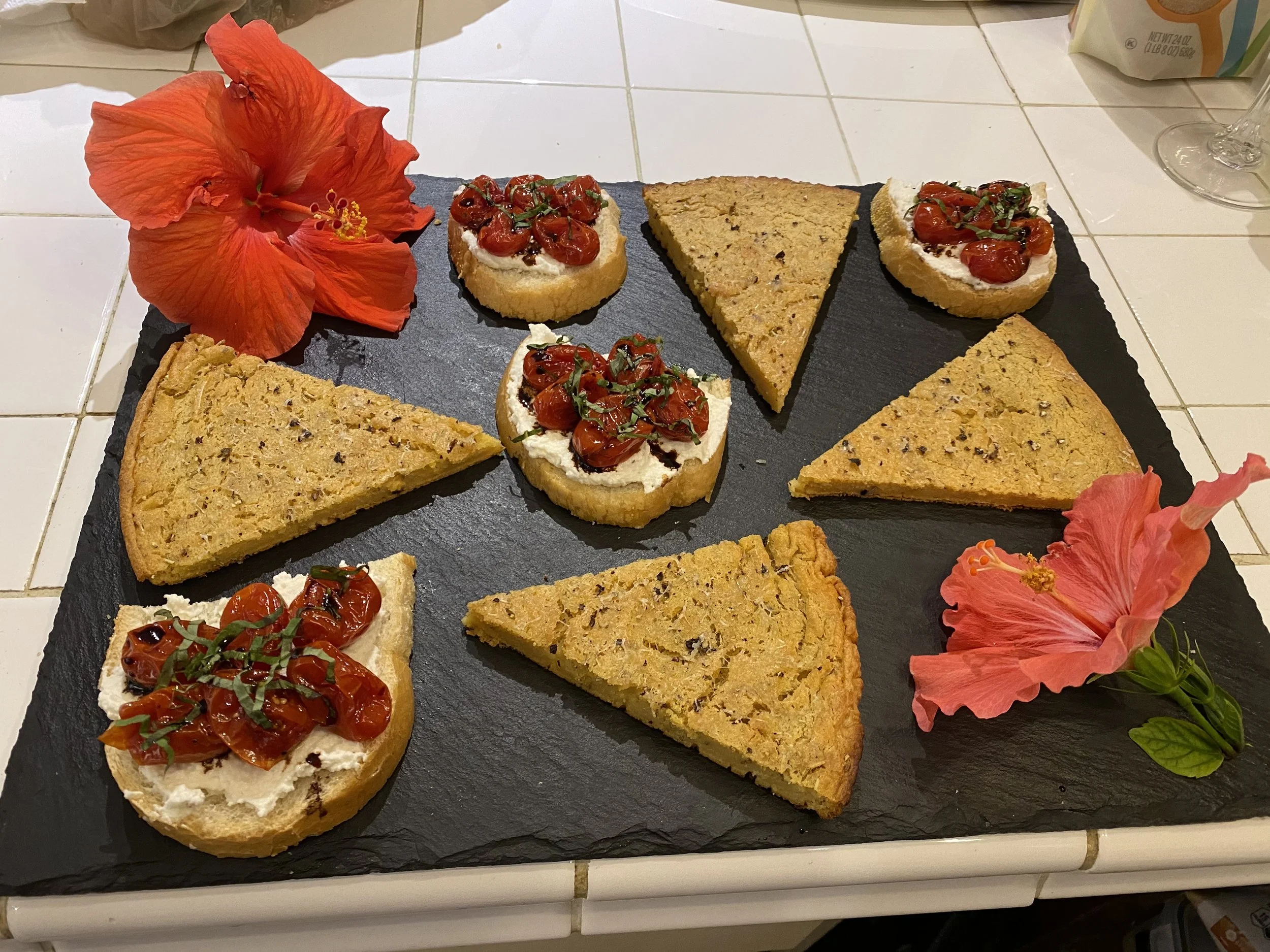 A black slate serving tray with slices of garlic bread and pieces of focaccia topped with cherry tomatoes and herbs, garnished with red hibiscus flowers.