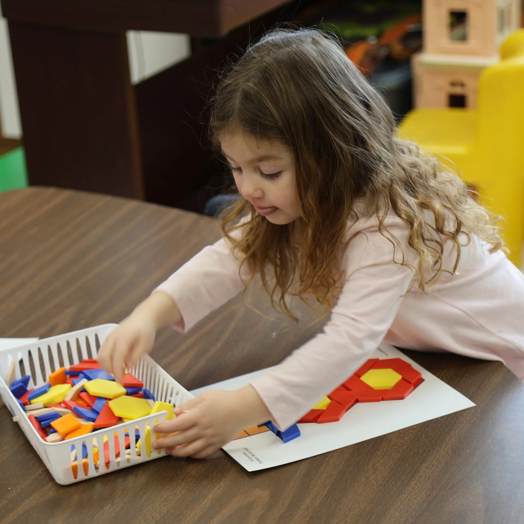 A young girl with curly brown hair wearing a pink long-sleeve shirt playing with colorful geometric shapes in a white basket and on a white sheet of paper on a brown table in a classroom.