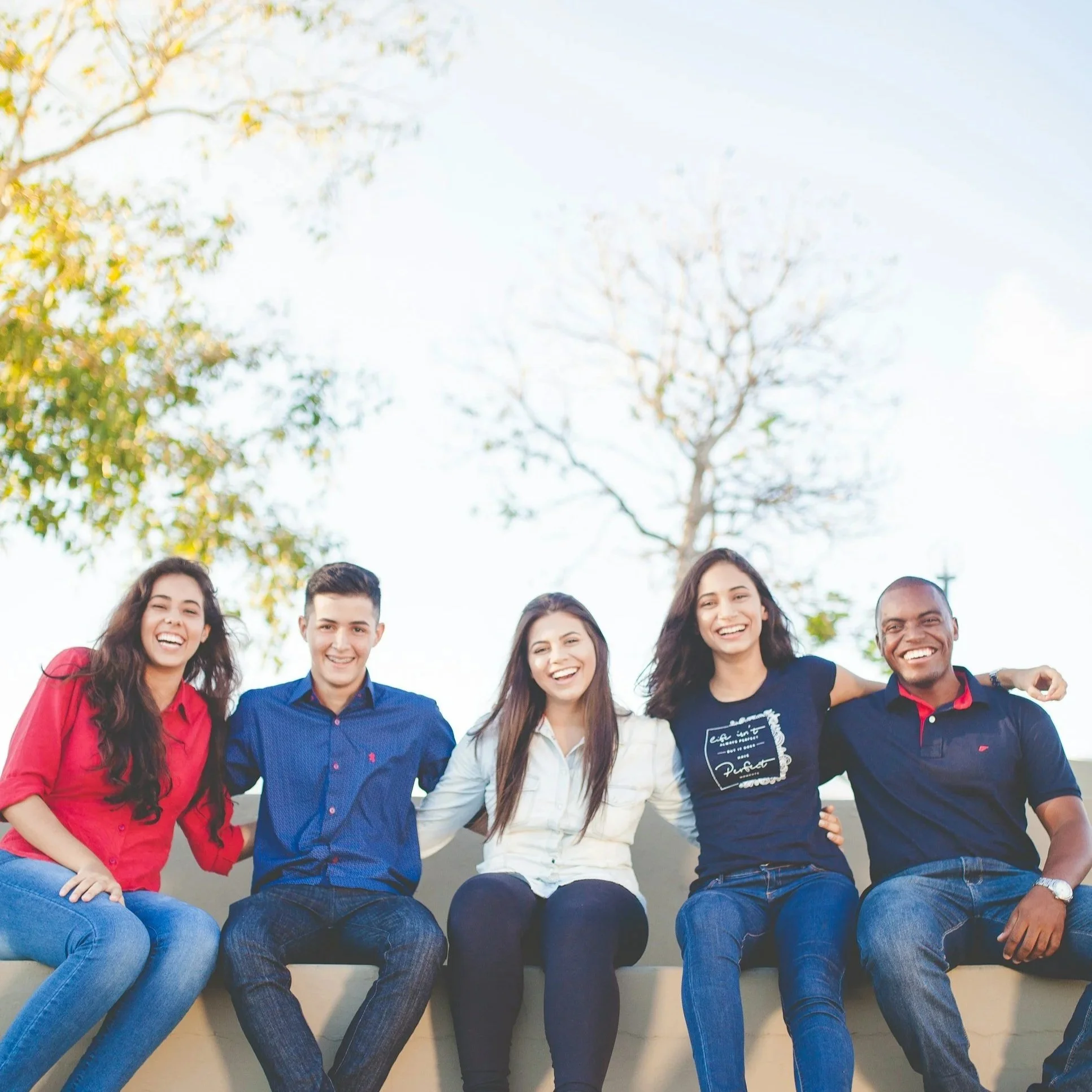 Group of five diverse young friends sitting outdoors on a bench, smiling, during a sunny day with trees in the background.