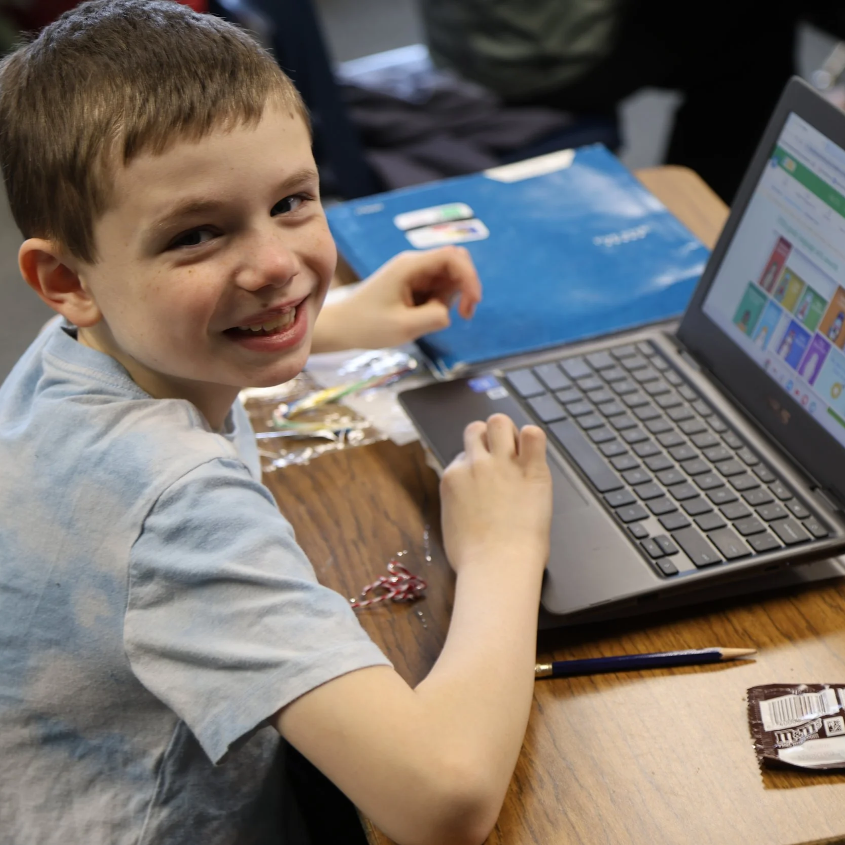A young boy with short brown hair and freckles sitting at a wooden desk using a laptop, smiling at the camera.