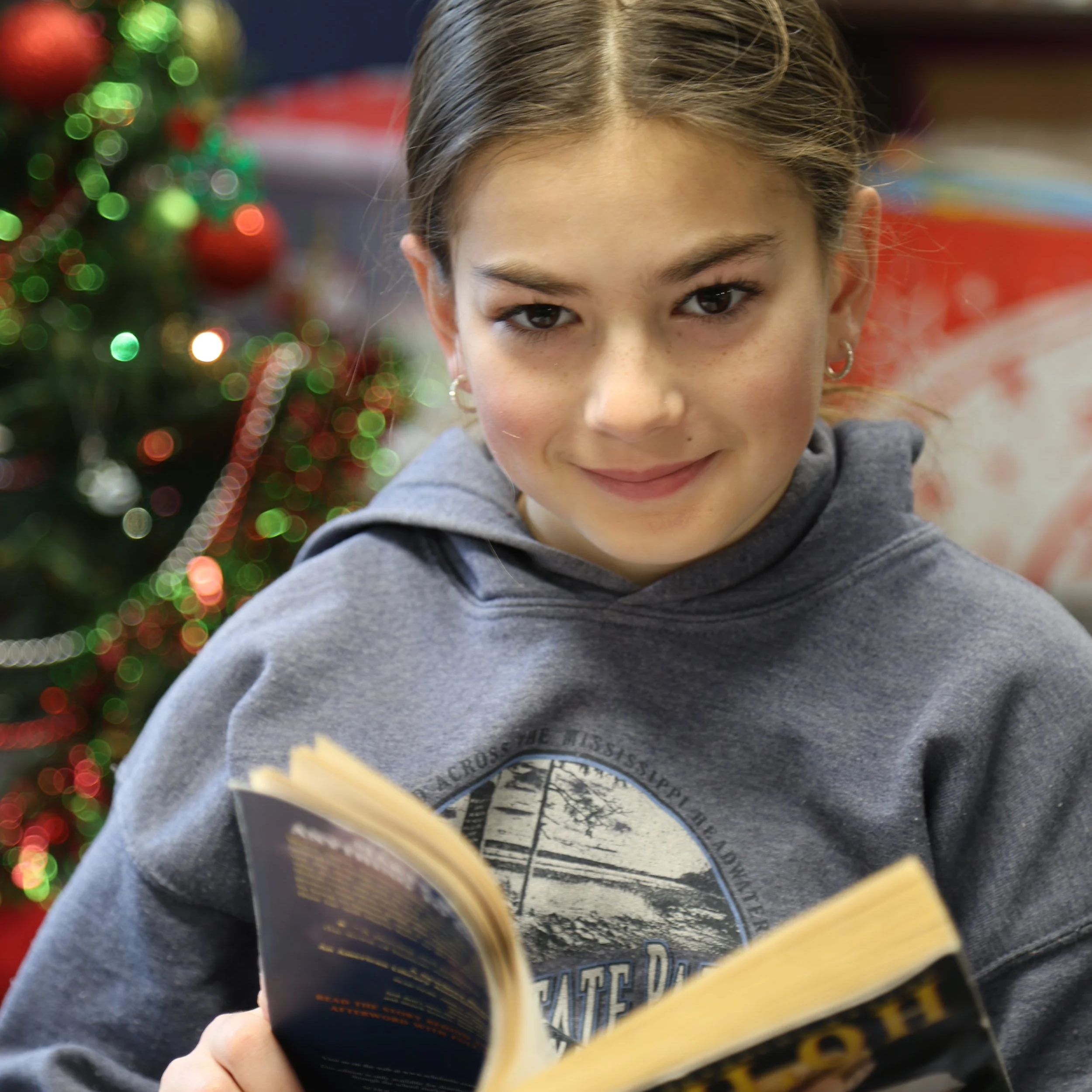 A young girl with brown hair in a braid, wearing hoop earrings and a gray hoodie, smiling while holding a book, with a blurred Christmas tree in the background.