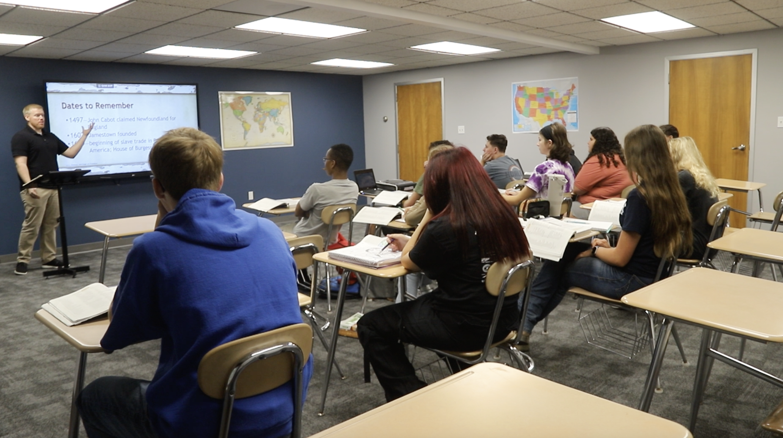 A classroom with students sitting at desks and listening to a teacher presenting at the front. The teacher is pointing at a large screen displaying a presentation titled 'Dates to Remember,' with maps of the world and the United States on the walls.