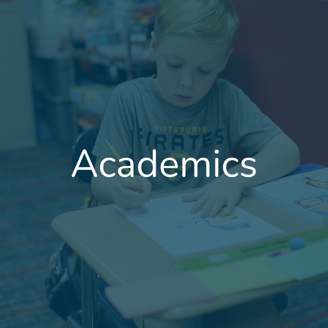 A young boy sitting at a table, drawing or coloring on a piece of paper in a classroom or library setting, with the word 'Academics' overlaid.