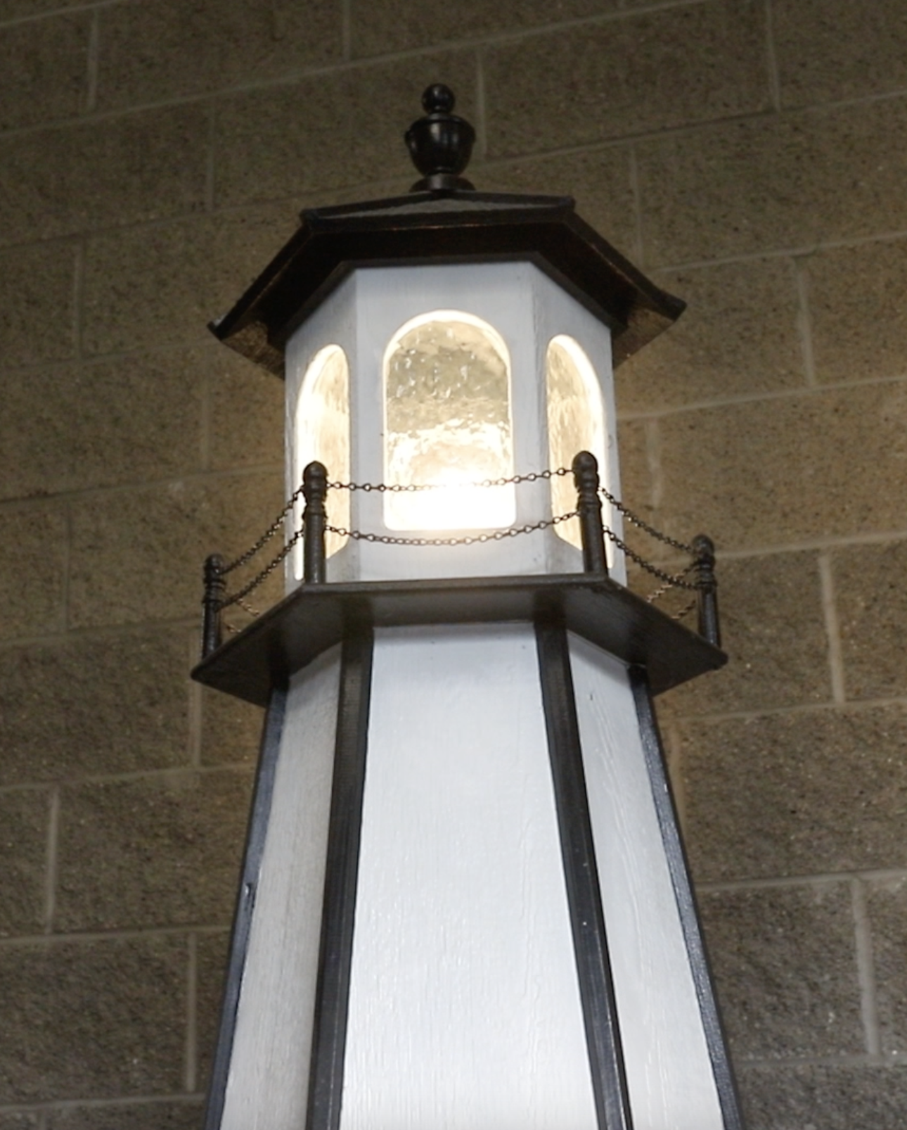 Close-up of a decorative outdoor lamp with a white paneled base, black accents, and a small balcony with chain railings, mounted on a brick wall.