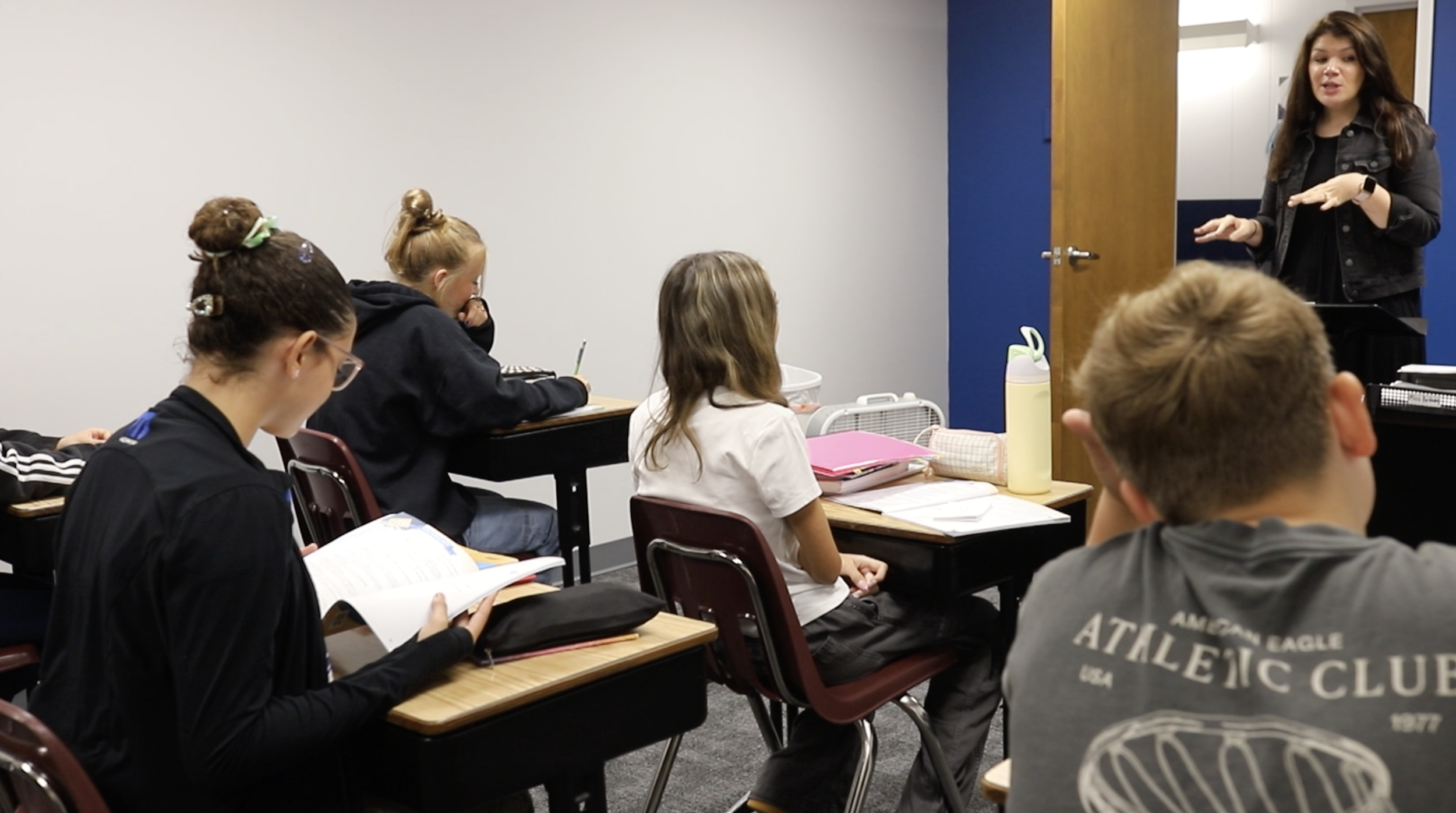 Teacher standing at the front of a classroom speaking to four students sitting at desks, with notebooks and textbooks open in front of them.