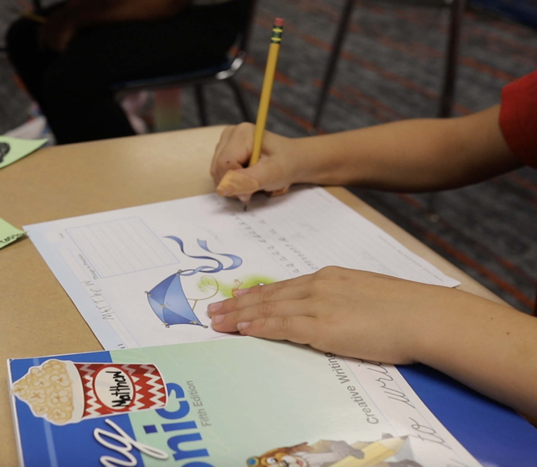 A person's hands are writing on a worksheet with a pencil, with a popcorn box nearby on a wooden table. The worksheet features a drawing of an umbrella, a tornado, and a lightning bolt, indicating a weather project or worksheet.