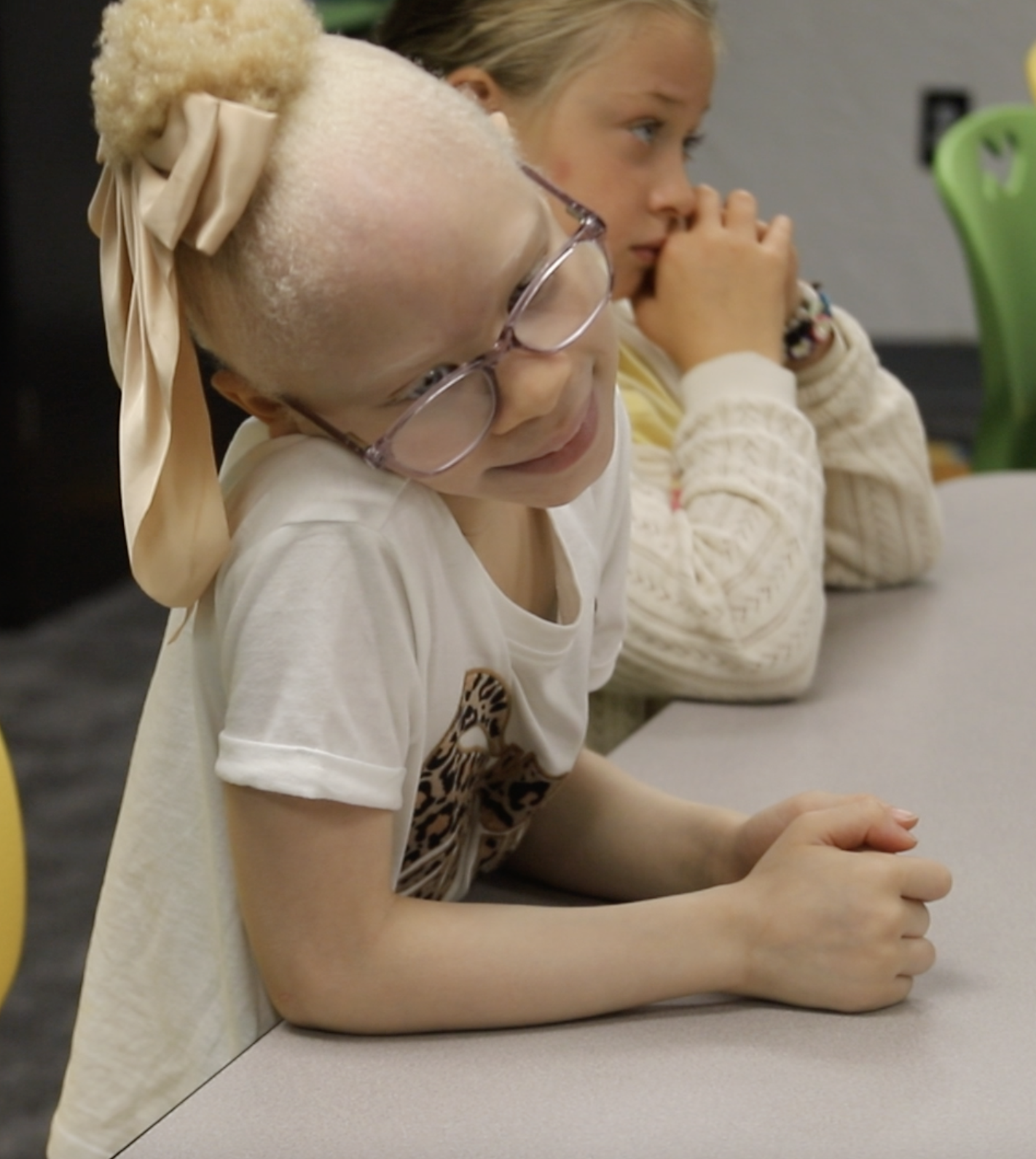 Two young girls sitting at a table, with one girl in the foreground wearing glasses and a beige bow in her hair, resting her arm on the table and smiling, the other girl in the background with her hands clasped near her face, both appear focused on something off-camera.