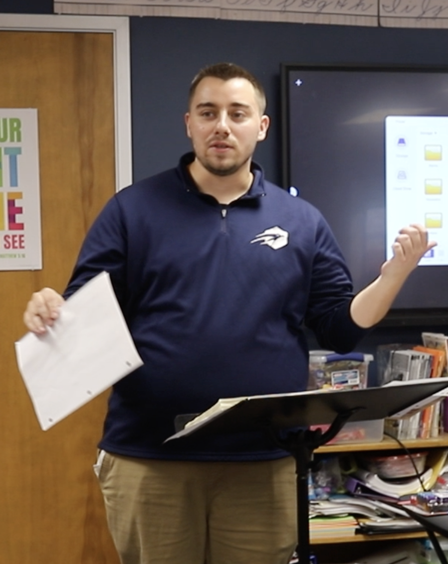 A man in a navy blue zip-up shirt with a logo, holding papers in one hand and gesturing with the other, speaking in a classroom or meeting room.