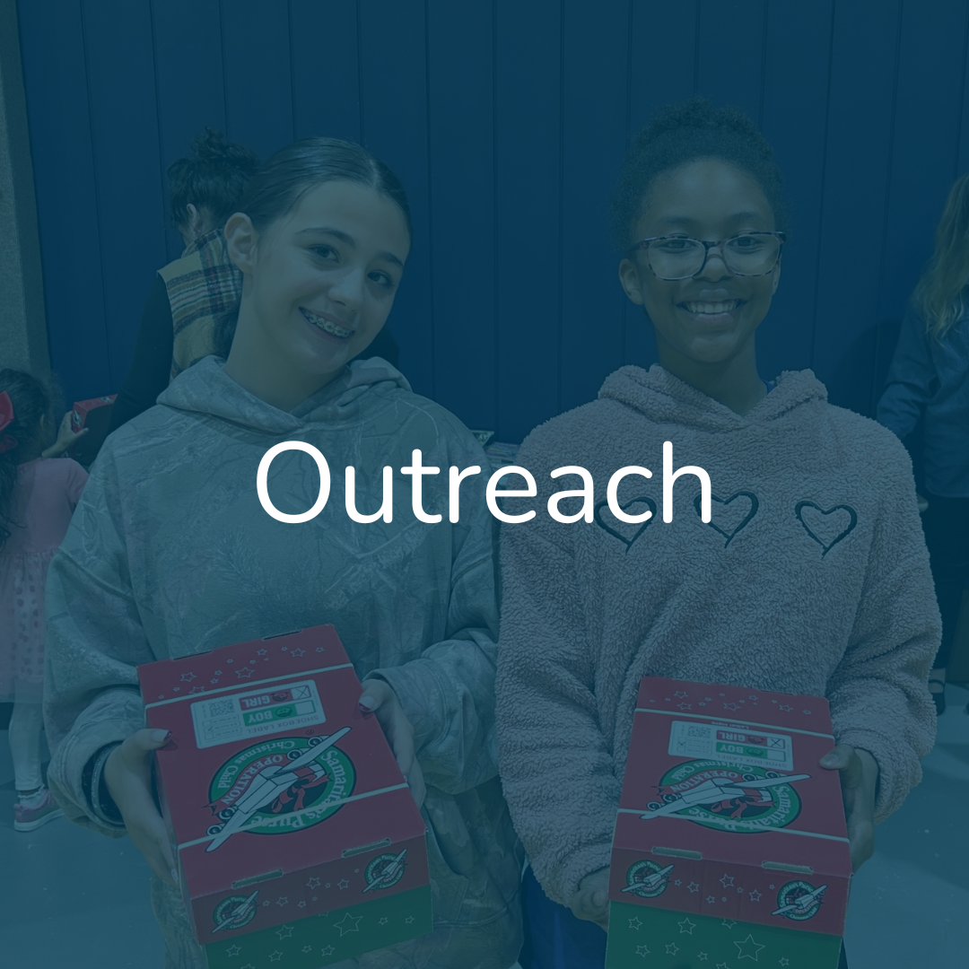 Two young girls smiling and holding pizza boxes, standing indoors with other children in the background, during outreach event.