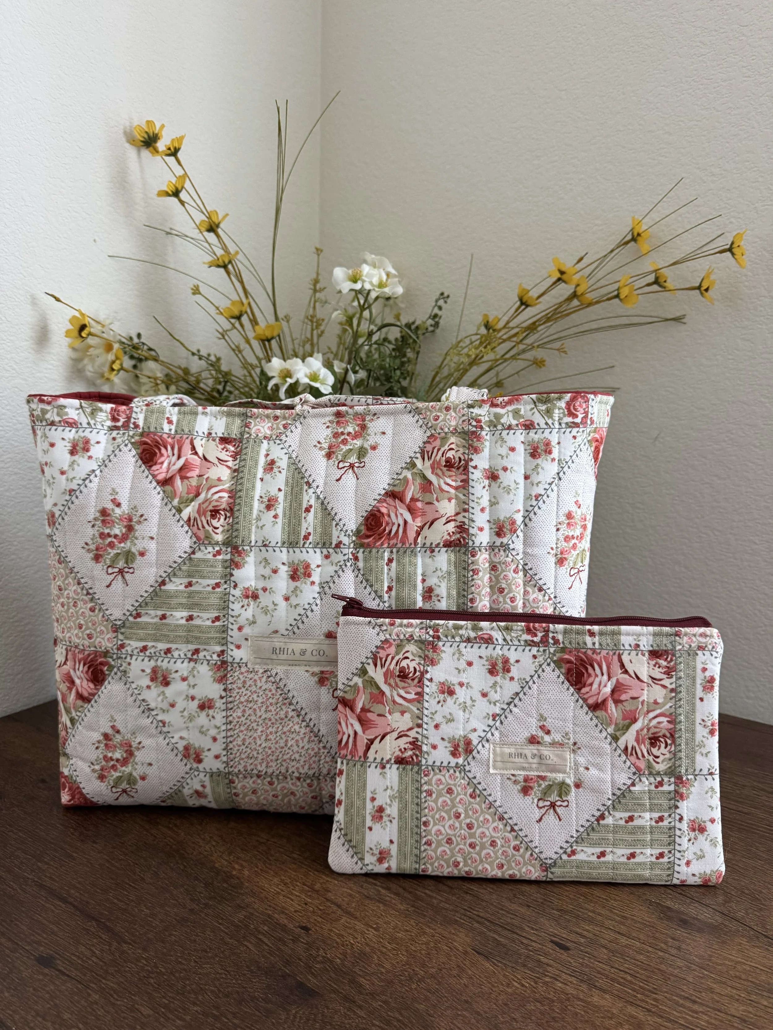A quilted fabric bag with a matching zippered pouch, both featuring a floral patchwork pattern of roses, stripes, and small dots, with a label 'RHIA & CO.' on each, placed on a wooden surface with a basket of yellow and white flowers in the background.