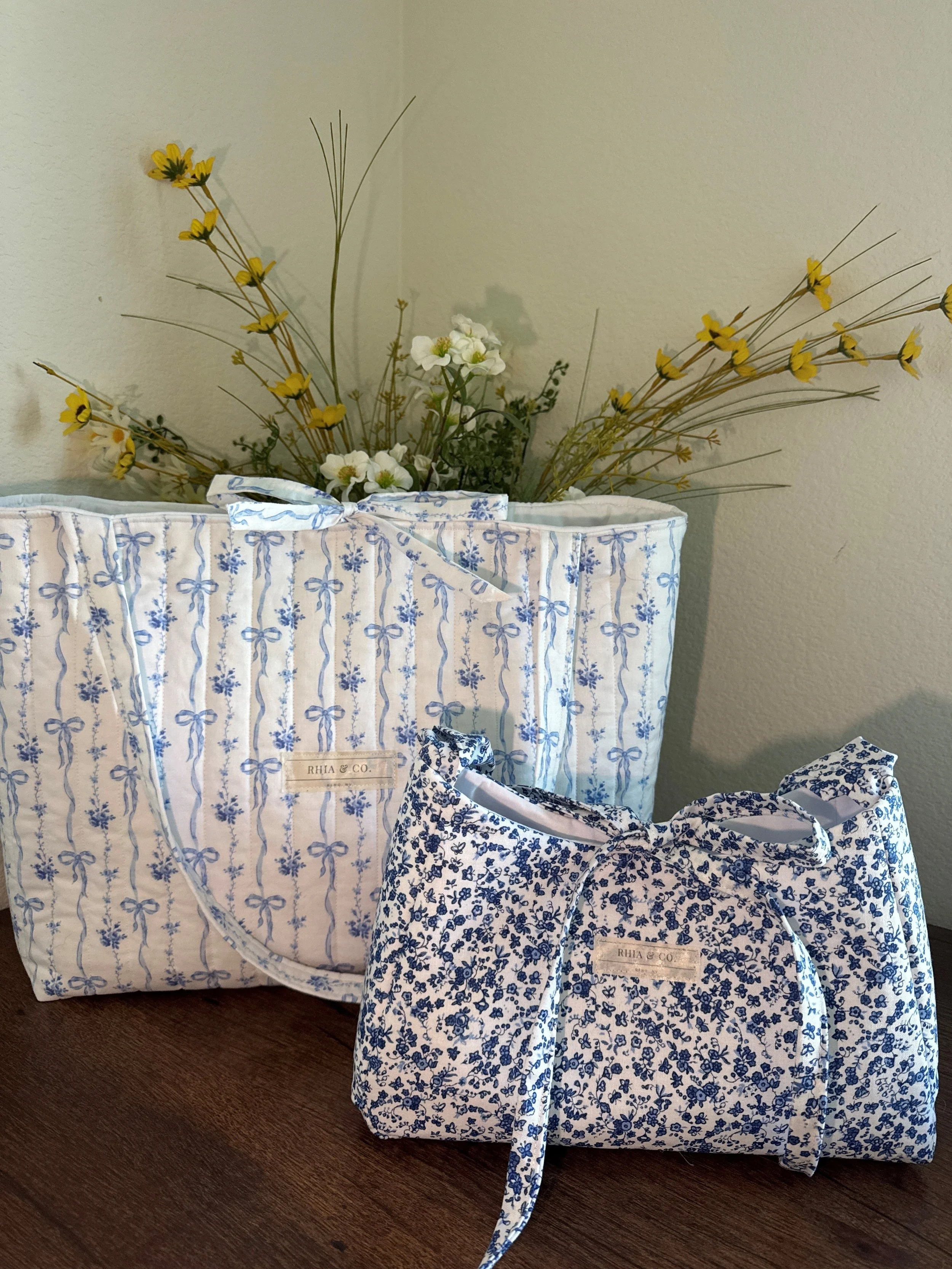 Two decorative fabric bags with blue floral patterns and ribbons, placed on a wooden surface in front of a beige wall with a floral arrangement in a fabric container.