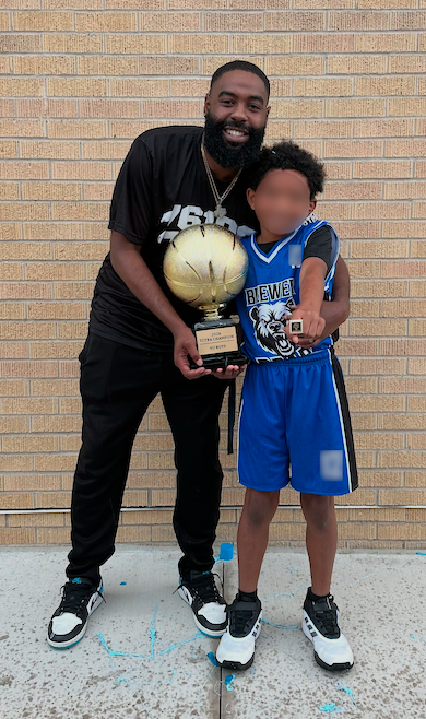 Coach Ross stands smiling beside a young player in a blue basketball uniform as they hold a championship trophy and ring outdoors.