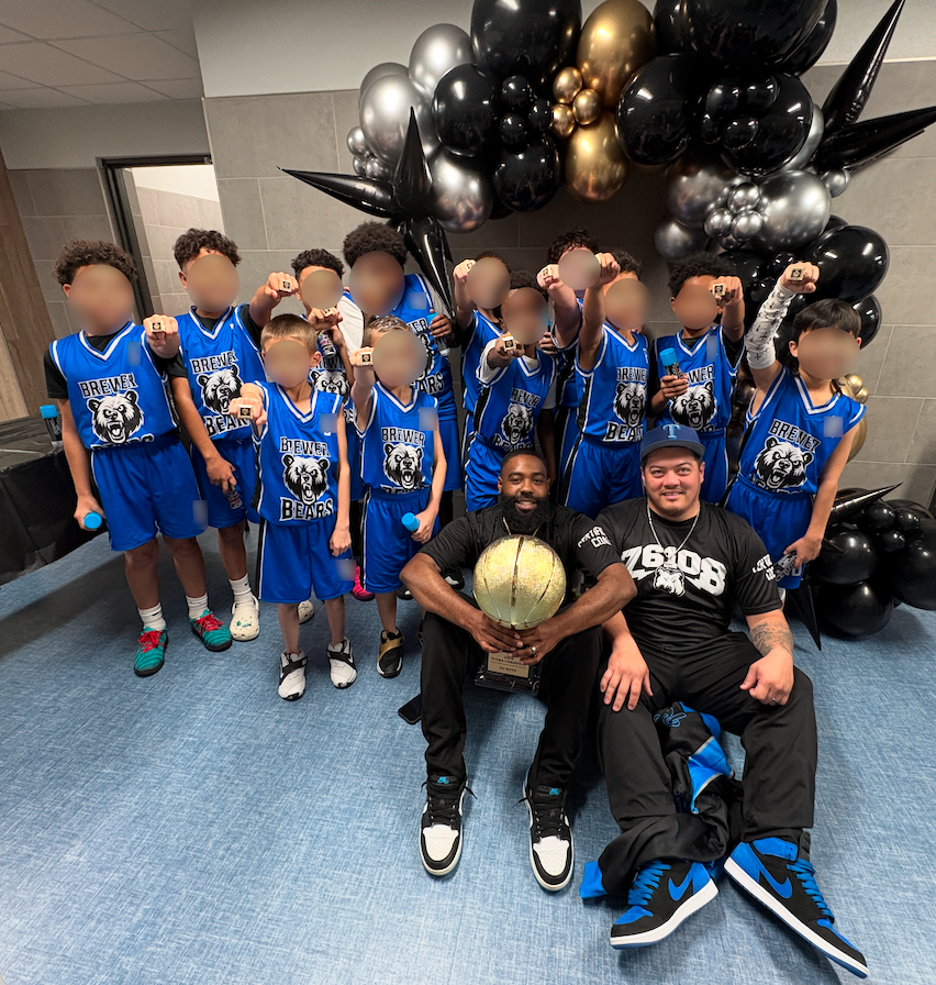 Coach Ross and assistant coach pose with the full youth basketball team, who are holding up championship rings in front of a black, silver, and gold balloon display.