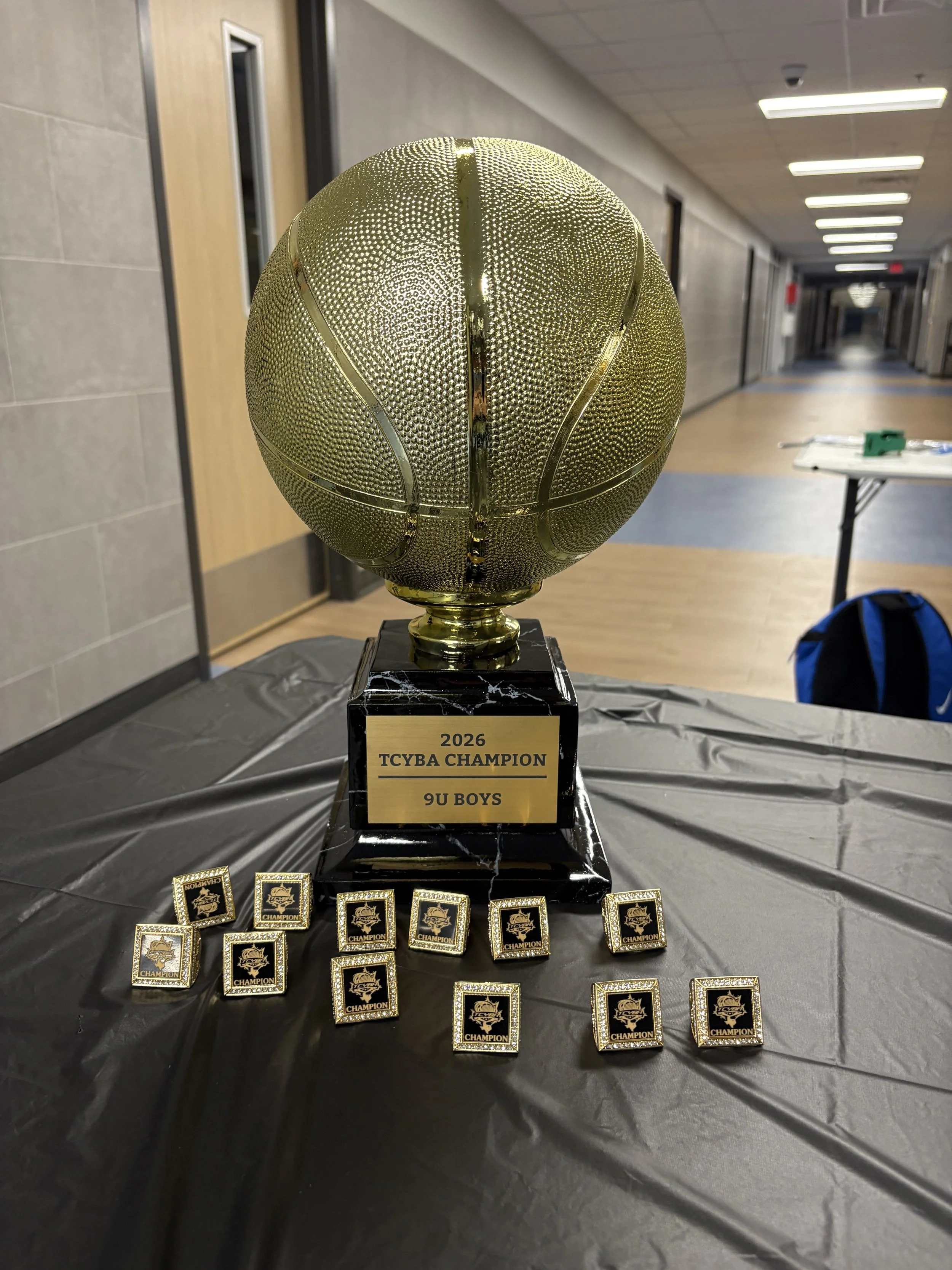 A gold basketball championship trophy sits on a table surrounded by individual championship rings for a 9U boys team.