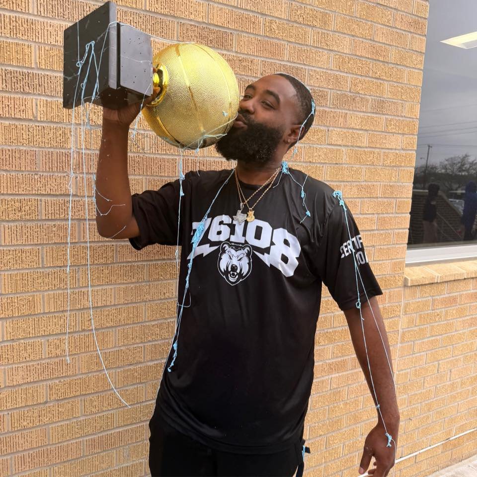 Coach Ross stands outdoors holding and kissing a gold basketball championship trophy while wearing team apparel after the season win.