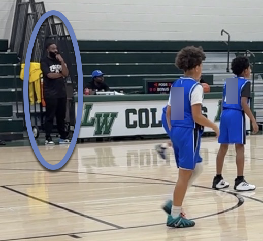 Coach Ross stands calmly on the sideline during a youth basketball game, watching the court while players in blue uniforms move in the foreground.