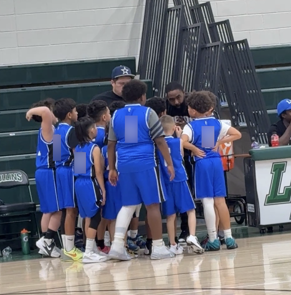 Coach Ross leans down into a close huddle with his team while an assistant coach stands nearby and the boys listen closely.