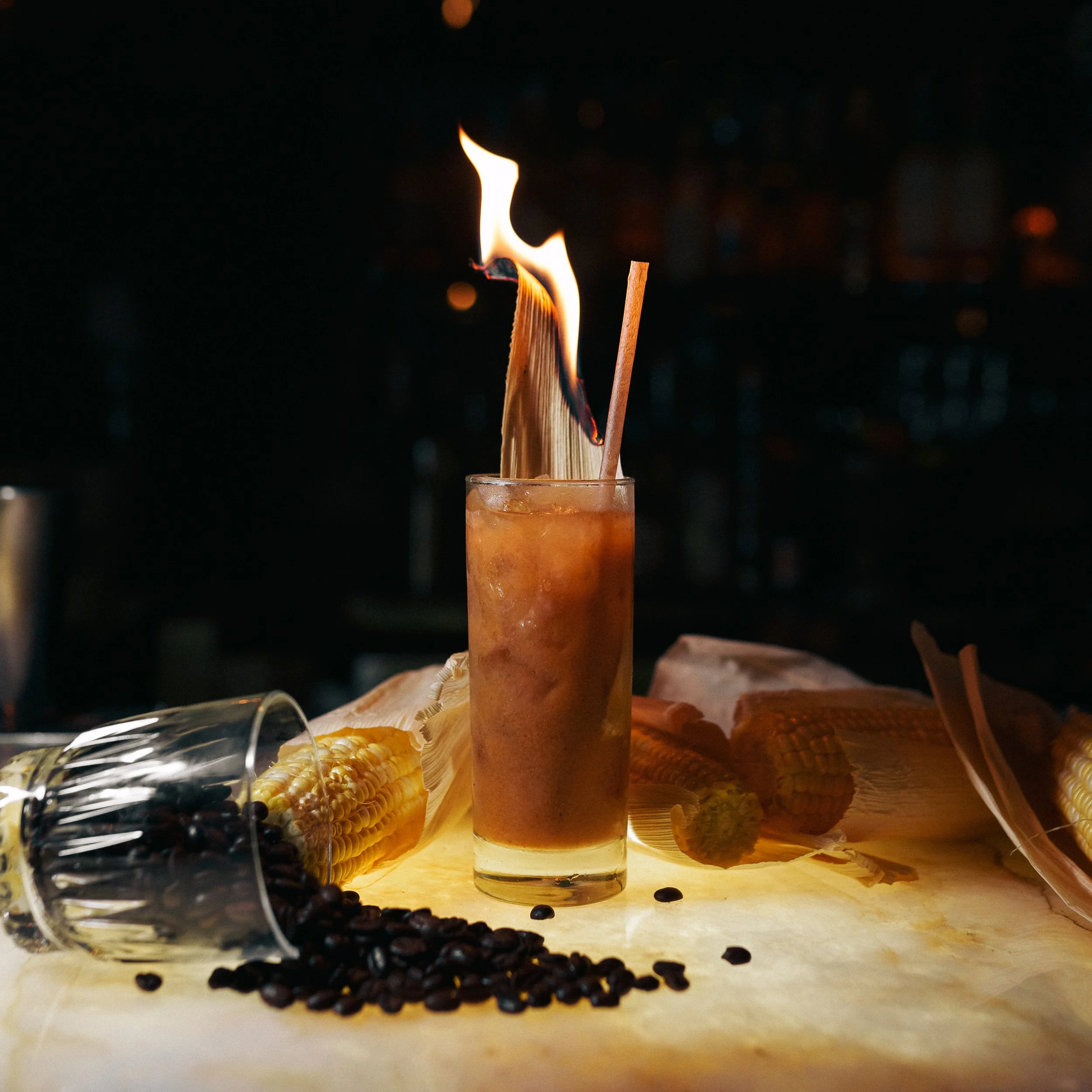A cocktail with corn on the cob and fire on top, surrounded by corn and coffee beans.