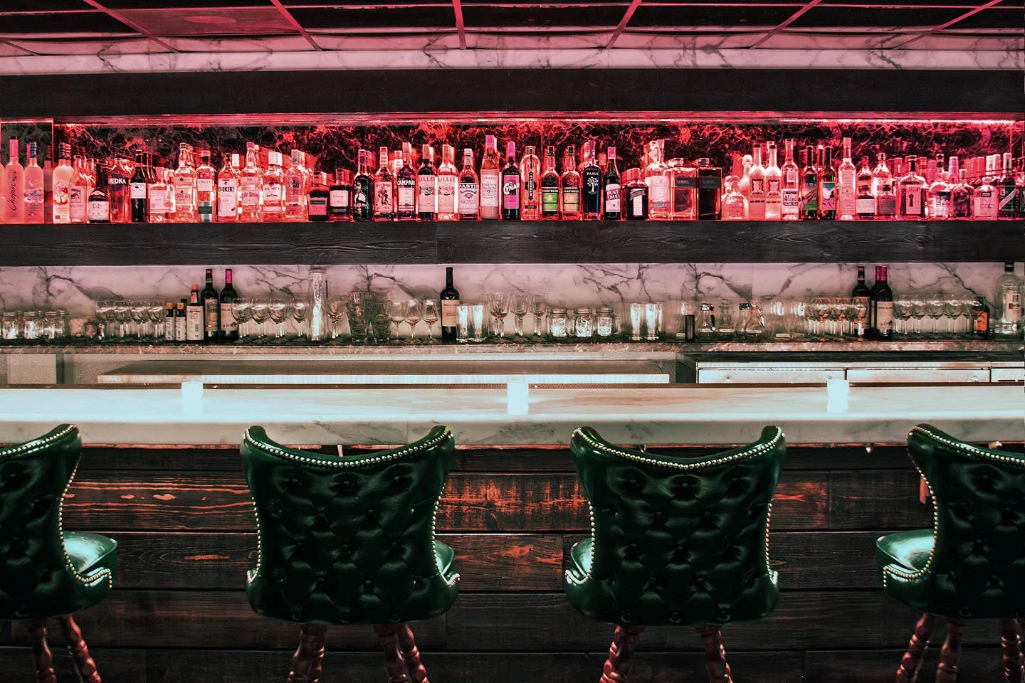 Bar with a marble countertop, shelves of various liquor bottles illuminated with pink and white lights, and green leather bar stools with button-tufted backs.
