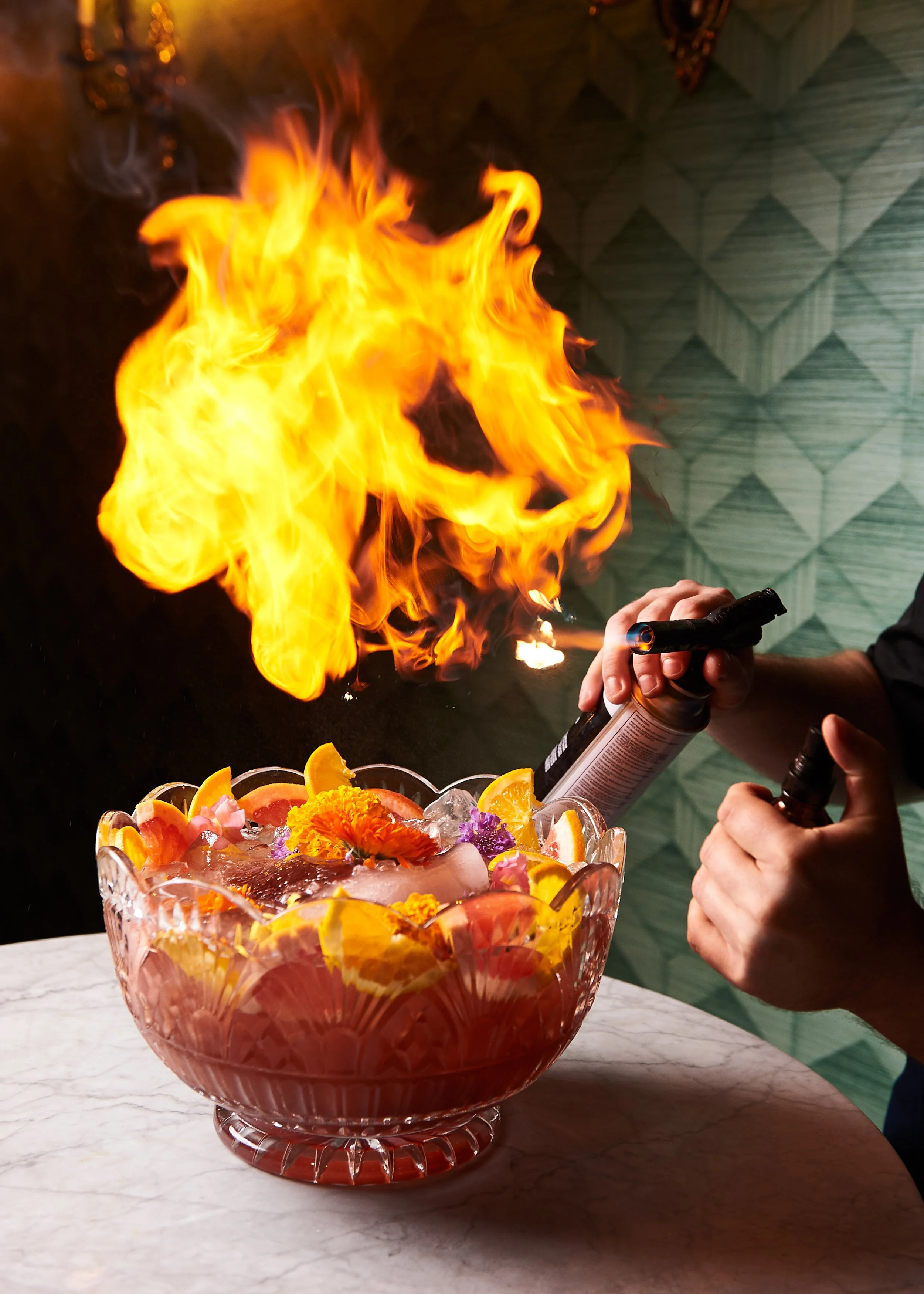 A bartender igniting a flaming substance over a bowl of colorful fruit punch garnished with oranges, flowers, and ice.