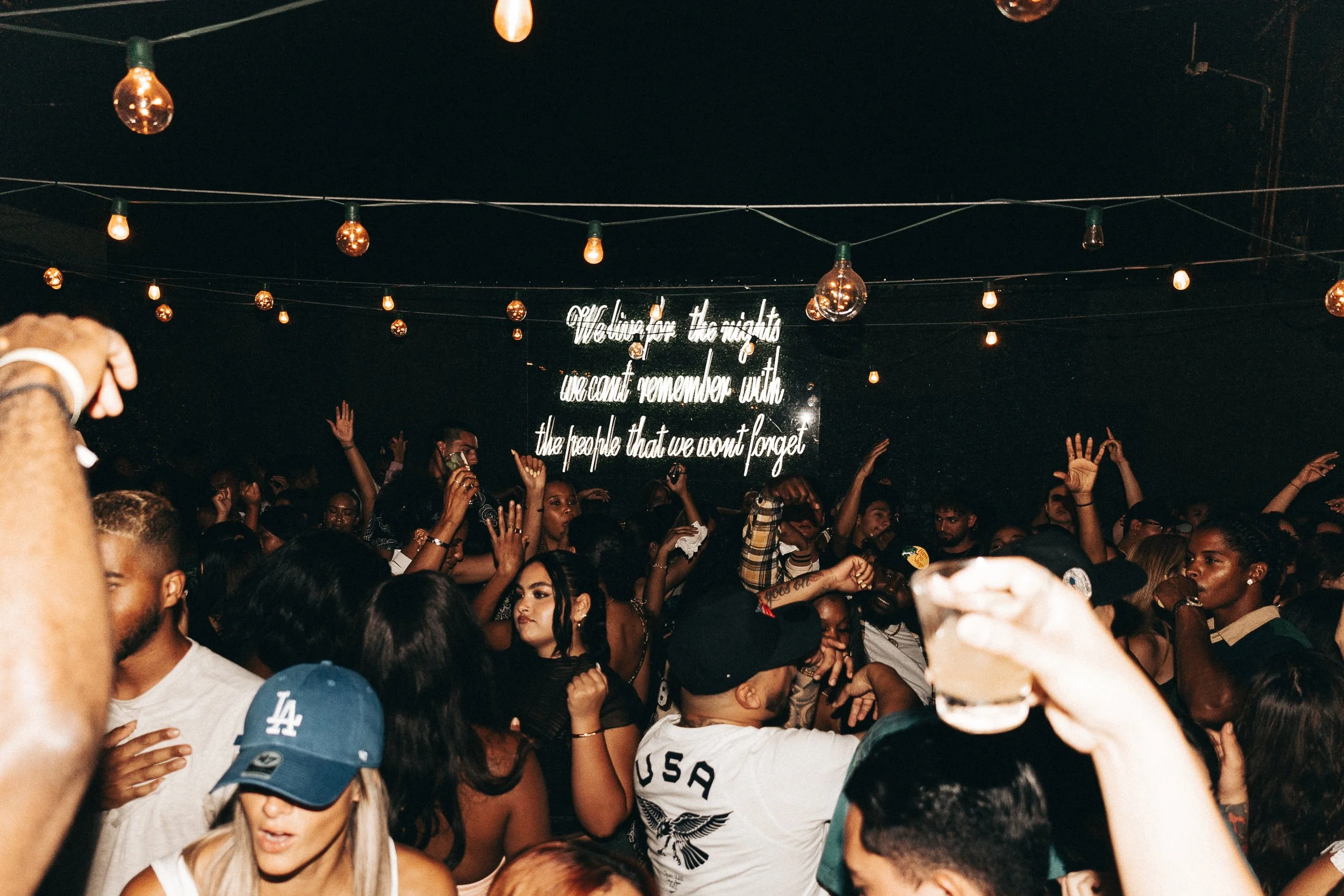Crowd of people at a night event with string lights and a neon sign background