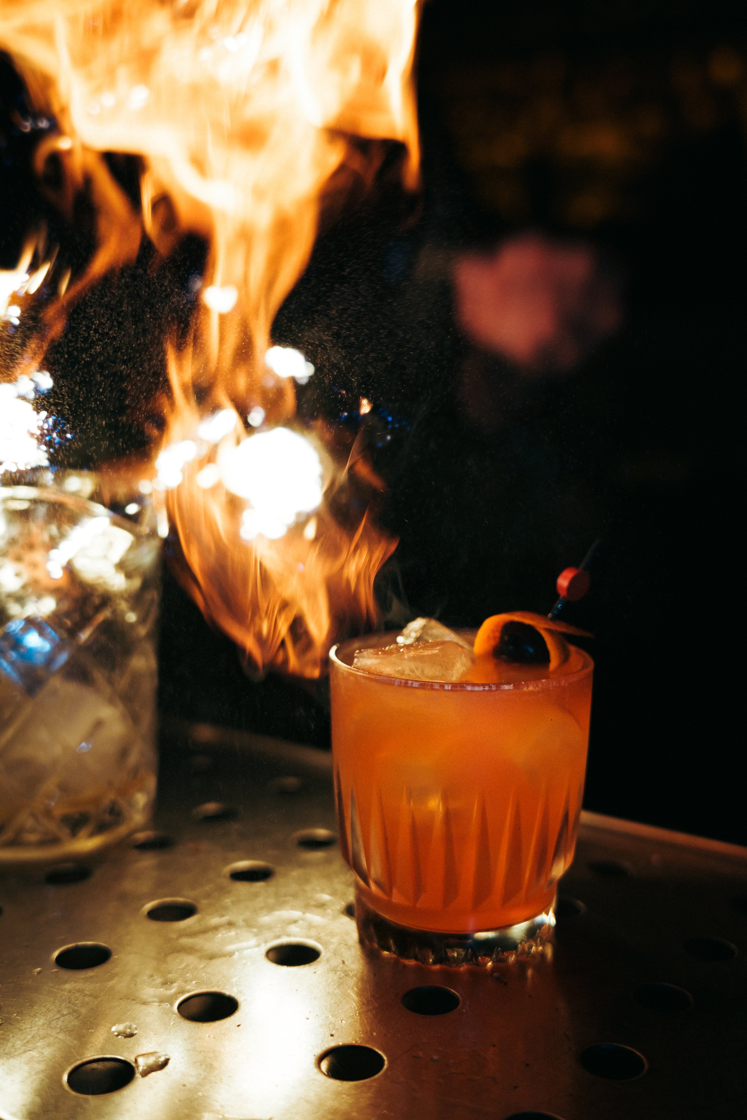 A cocktail with an orange hue and a cherry on a cocktail pick, set on a perforated metal surface, with a large flame in the background.