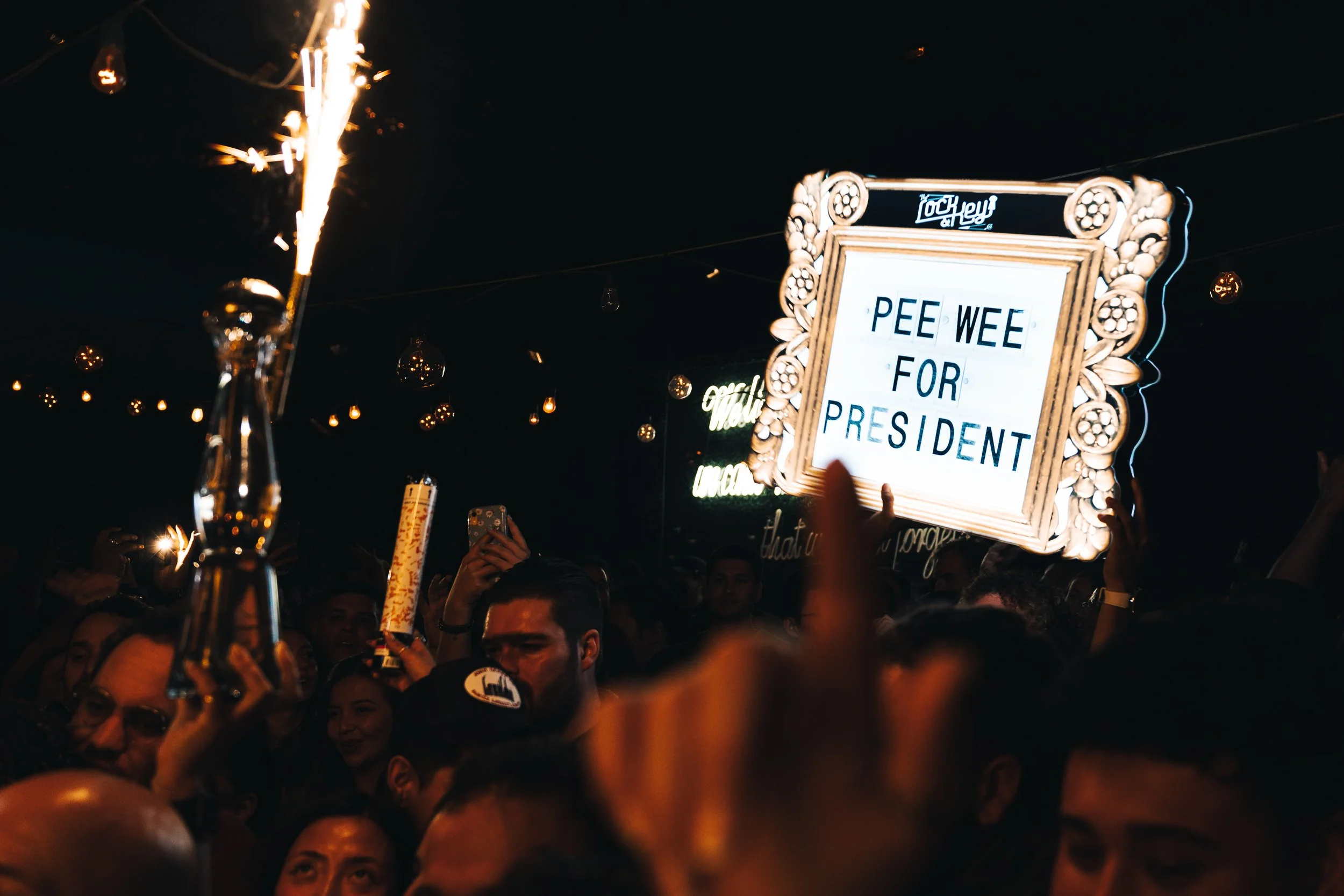 Crowd of people at a night event, some holding phones and signs. One sign reads 'PEE WEE FOR PRESIDENT' with decorative frame. String lights and neon signs are visible in the background, creating a lively atmosphere.