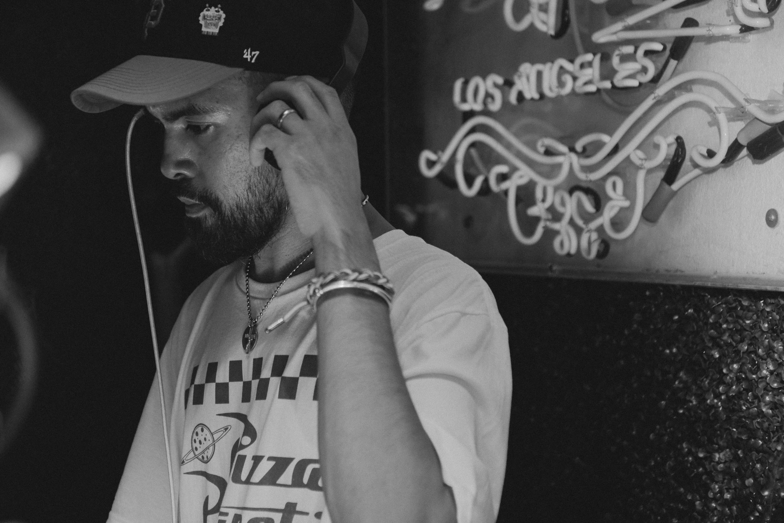A man with a beard wearing a cap and a t-shirt with a graphic, listening to music through earphones, standing against a wall with a neon sign that says 'Los Angeles'.
