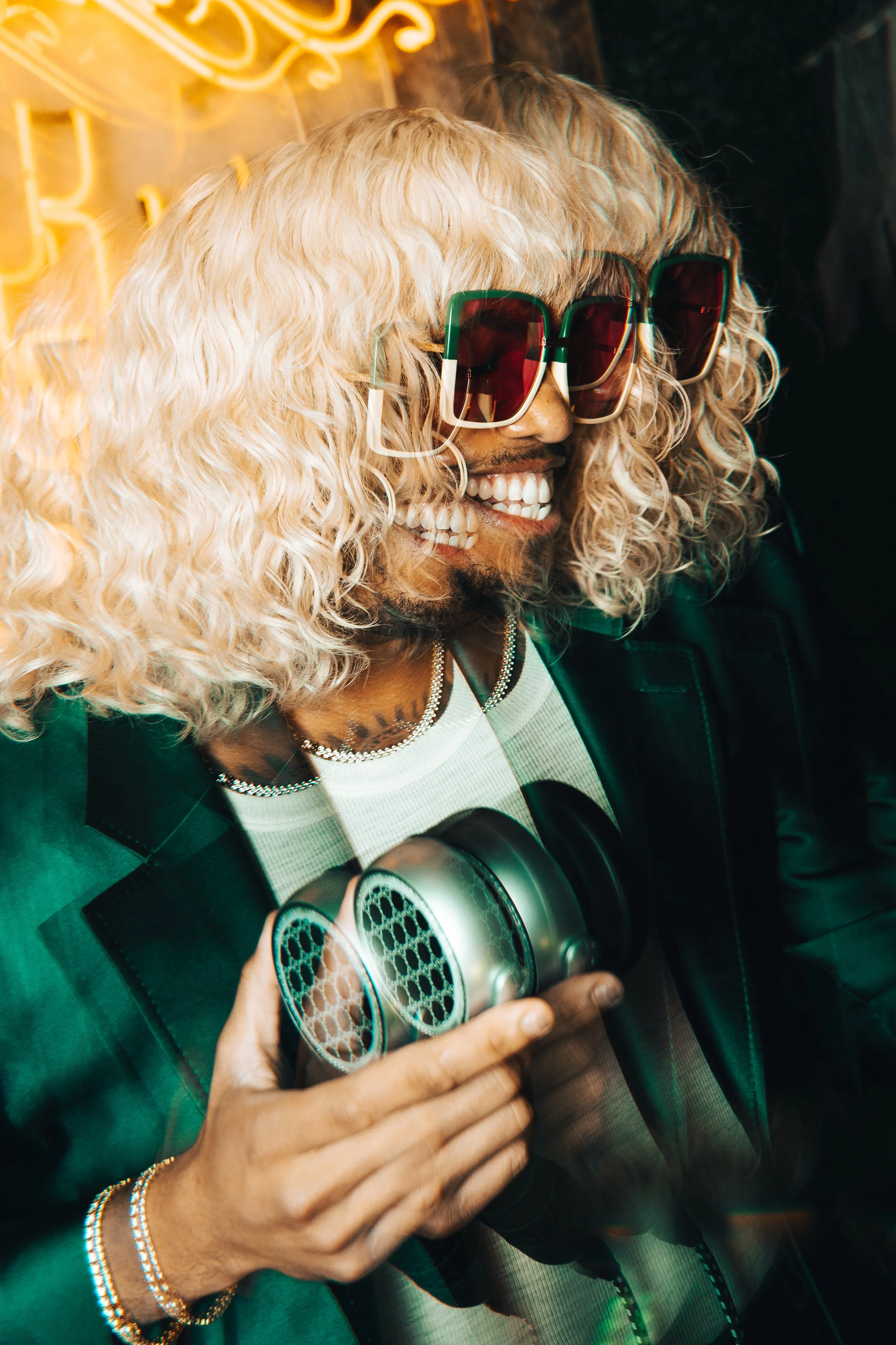 A person with curly platinum blonde hair, wearing large sunglasses with green frames, smiling, holding a metallic speaker, dressed in a black jacket and white shirt, standing in front of a neon light.
