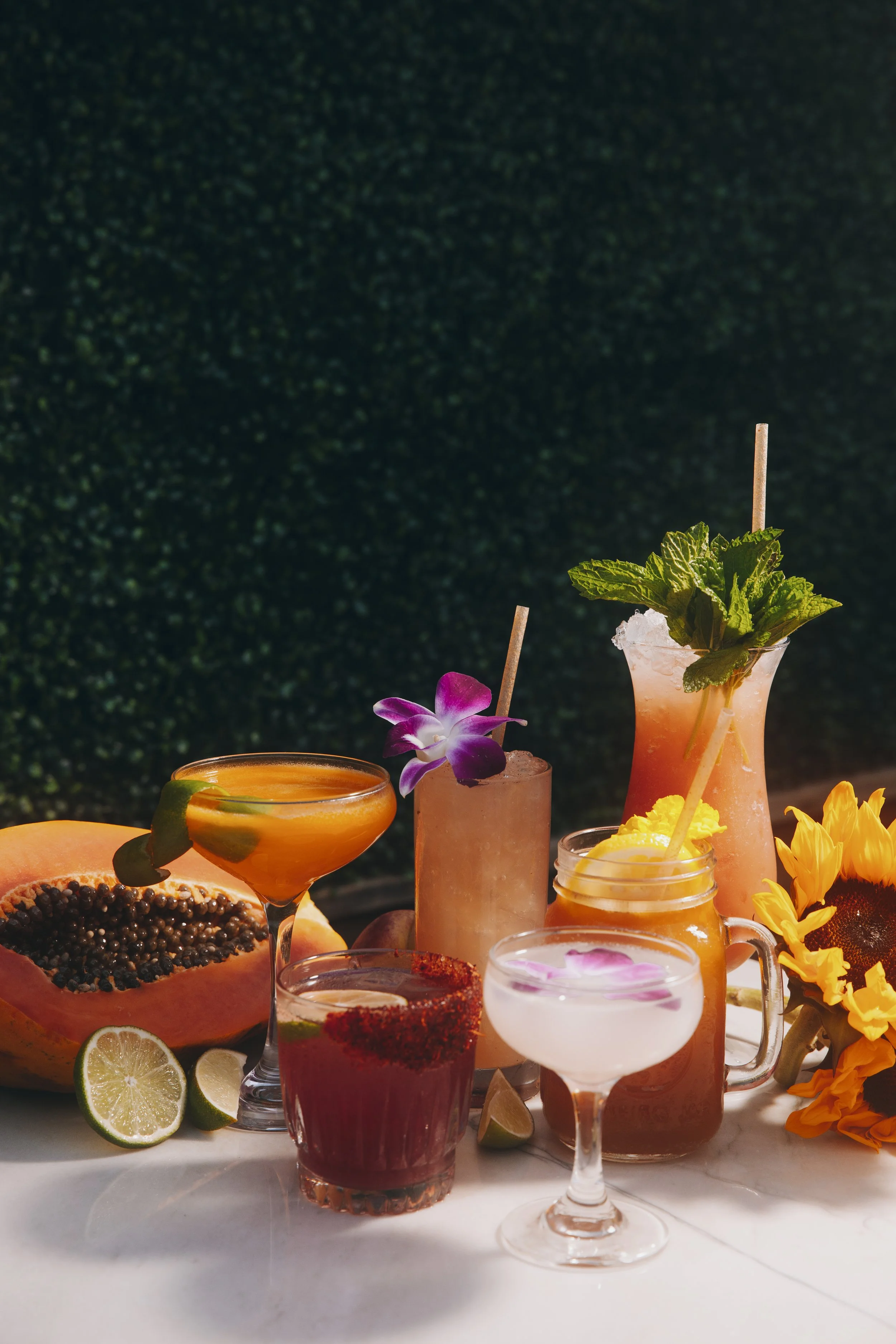 A variety of colorful cocktails on a white table, with fresh fruits and flowers, set against a dark background.