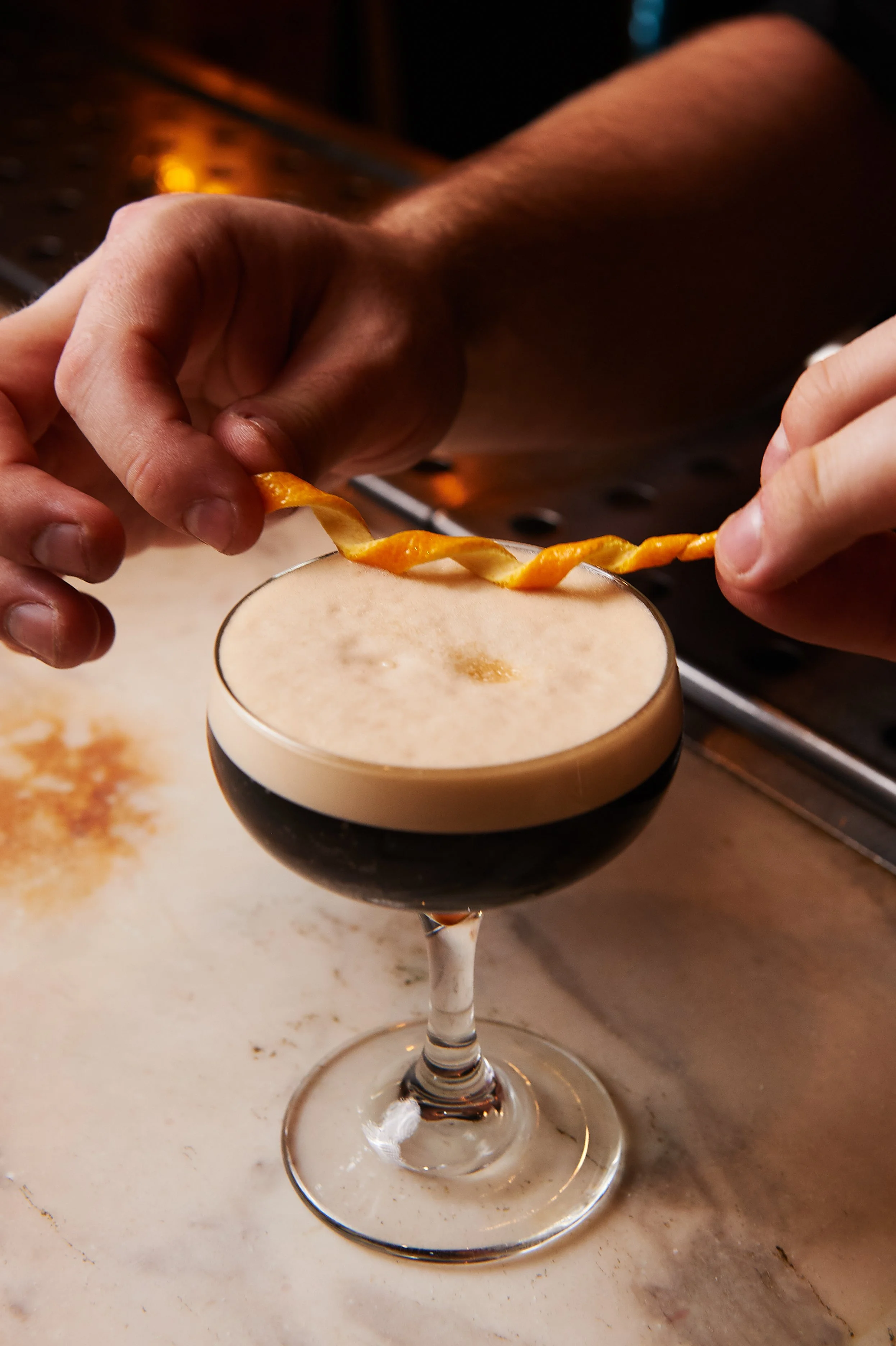 Person garnishing a dark cocktail with an orange twist twist.