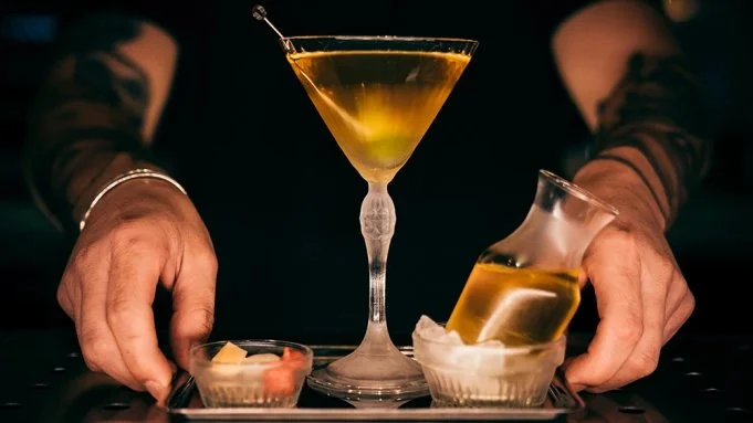 A bartender presents a tray with a martini glass and a pouring glass, both filled with a yellow cocktail. The bartender's hands are visible, placing the drinks on the tray.