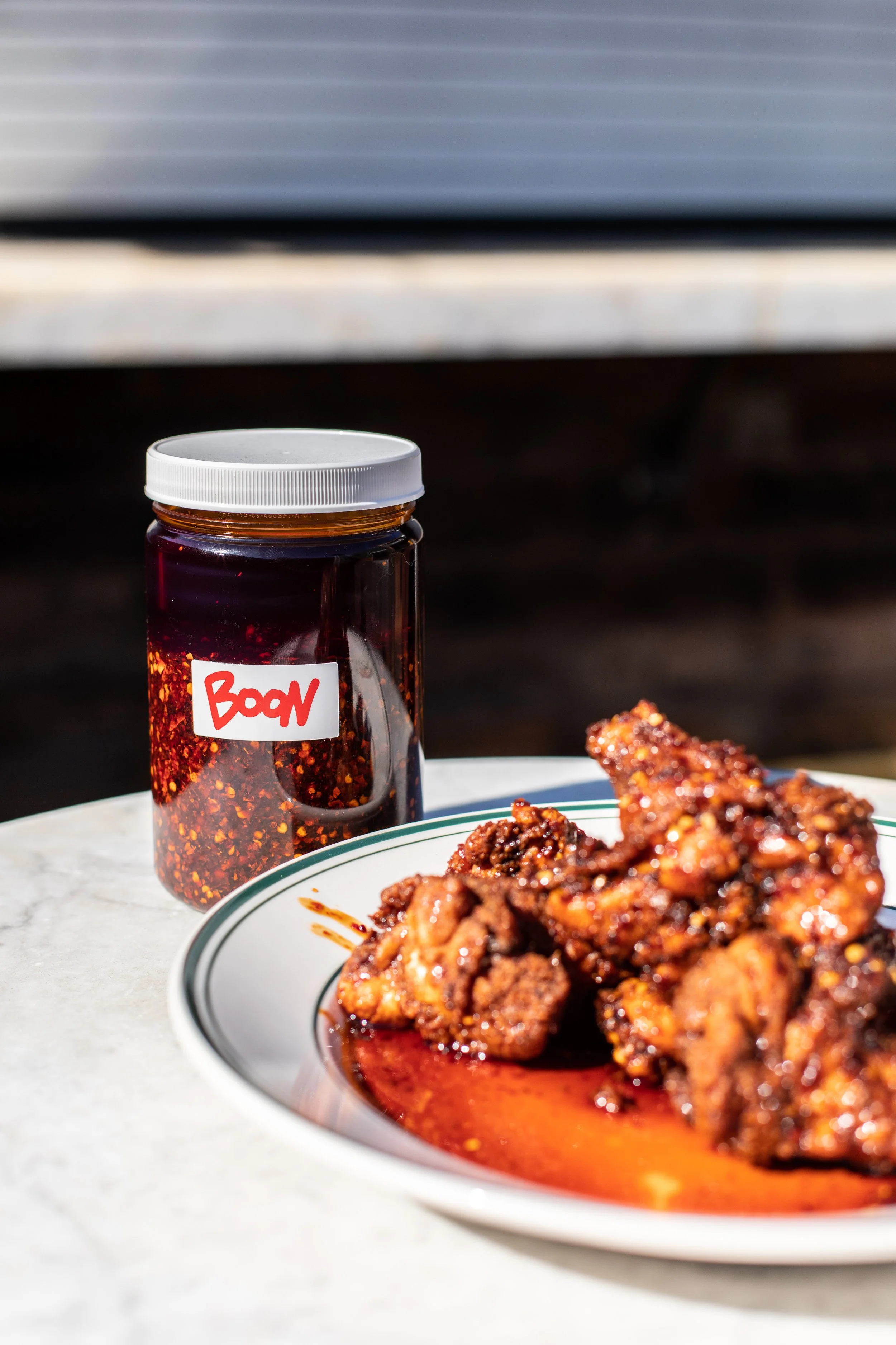 Plate of glazed fried chicken pieces and a jar of chili sauce labeled 'Boon' on a white table outside.
